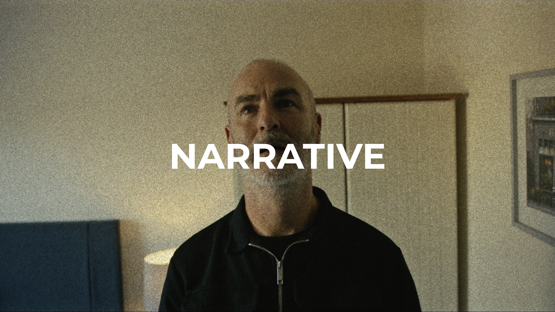 A bald man with a gray beard in a black jacket stands in a room with neutral-colored walls, a painting, and a lamp in the background, with the word 'NARRATIVE' overlaid on his face.