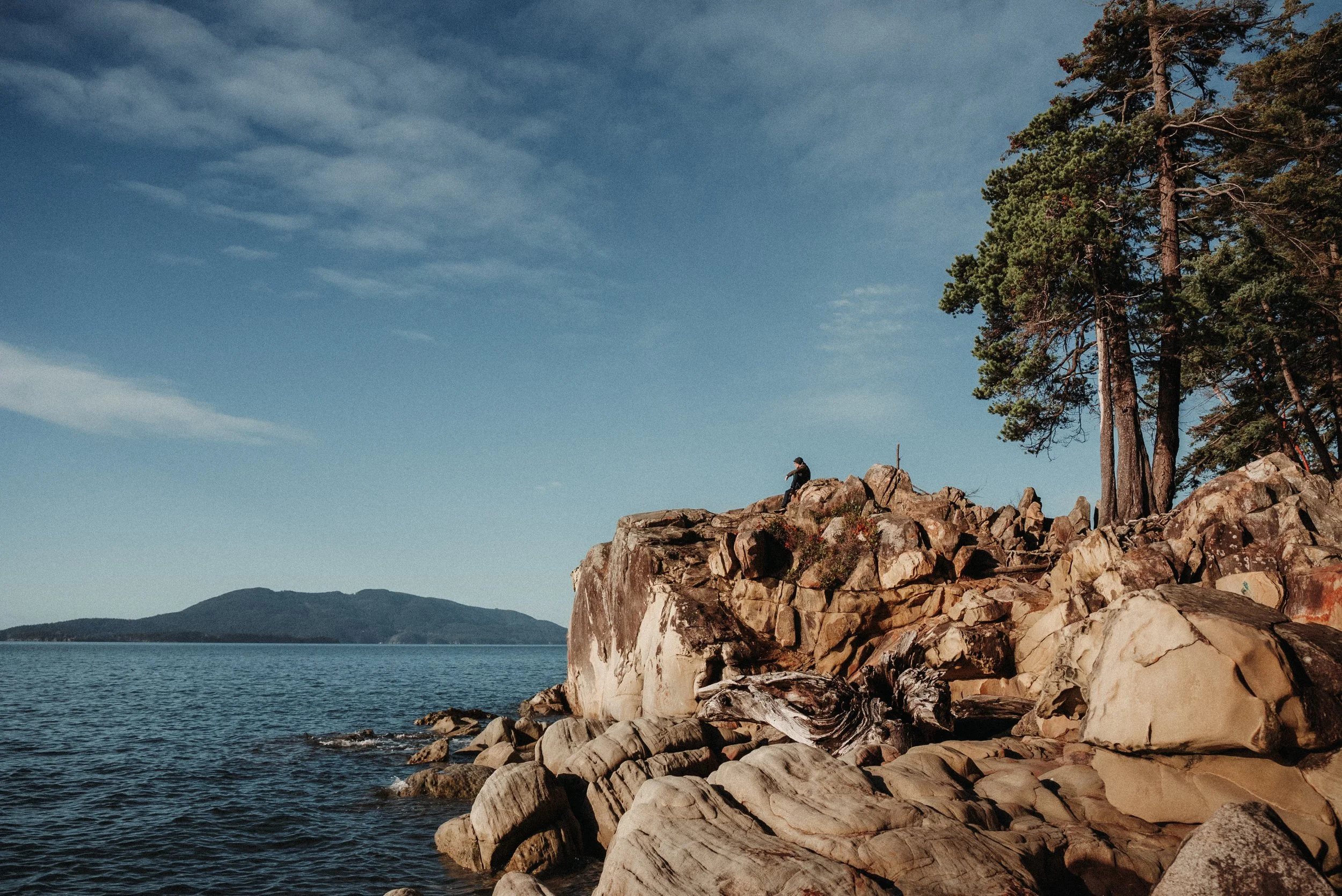 A person sitting on rocks near a lake with mountains in the background and pine trees to the right, under a partly cloudy sky.