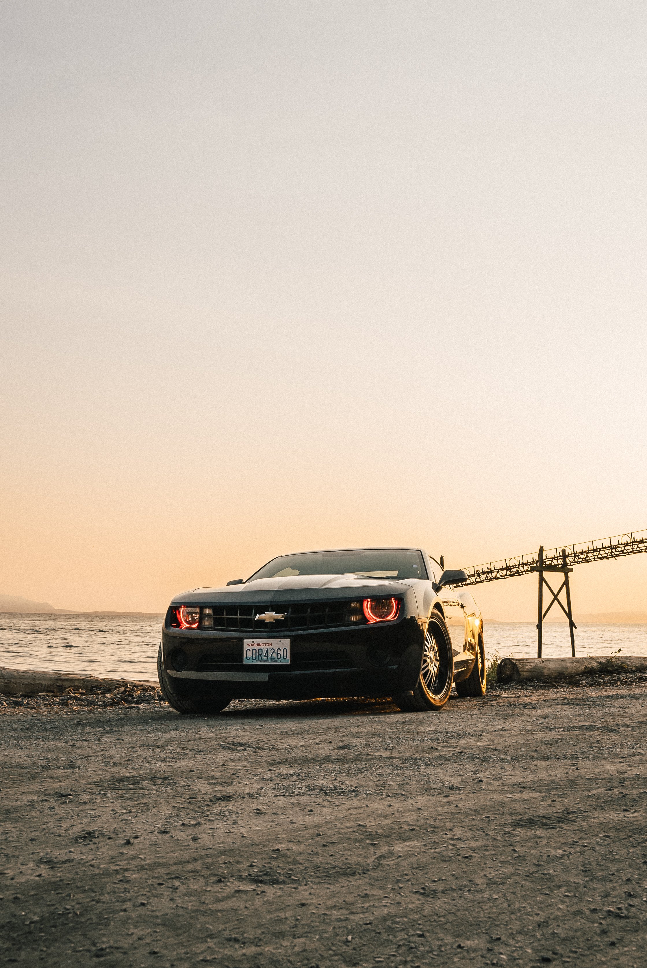 A black Chevrolet Camaro parked on a dirt surface near a body of water during sunset, with a bridge in the background.