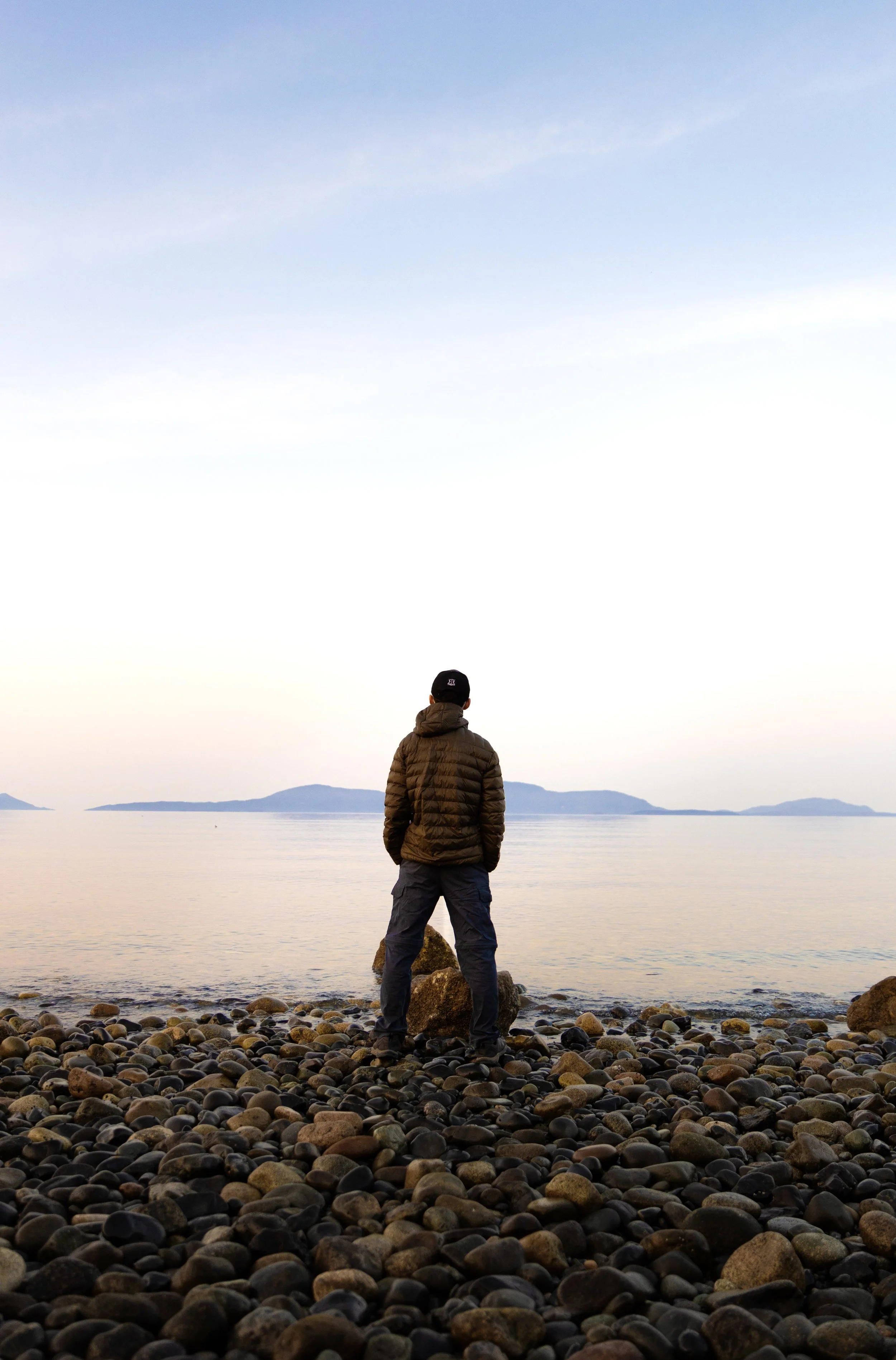 A person wearing a brown puffer jacket and a black cap stands on a rocky beach, looking out over a calm body of water with distant islands under a partly cloudy sky at dusk or dawn.