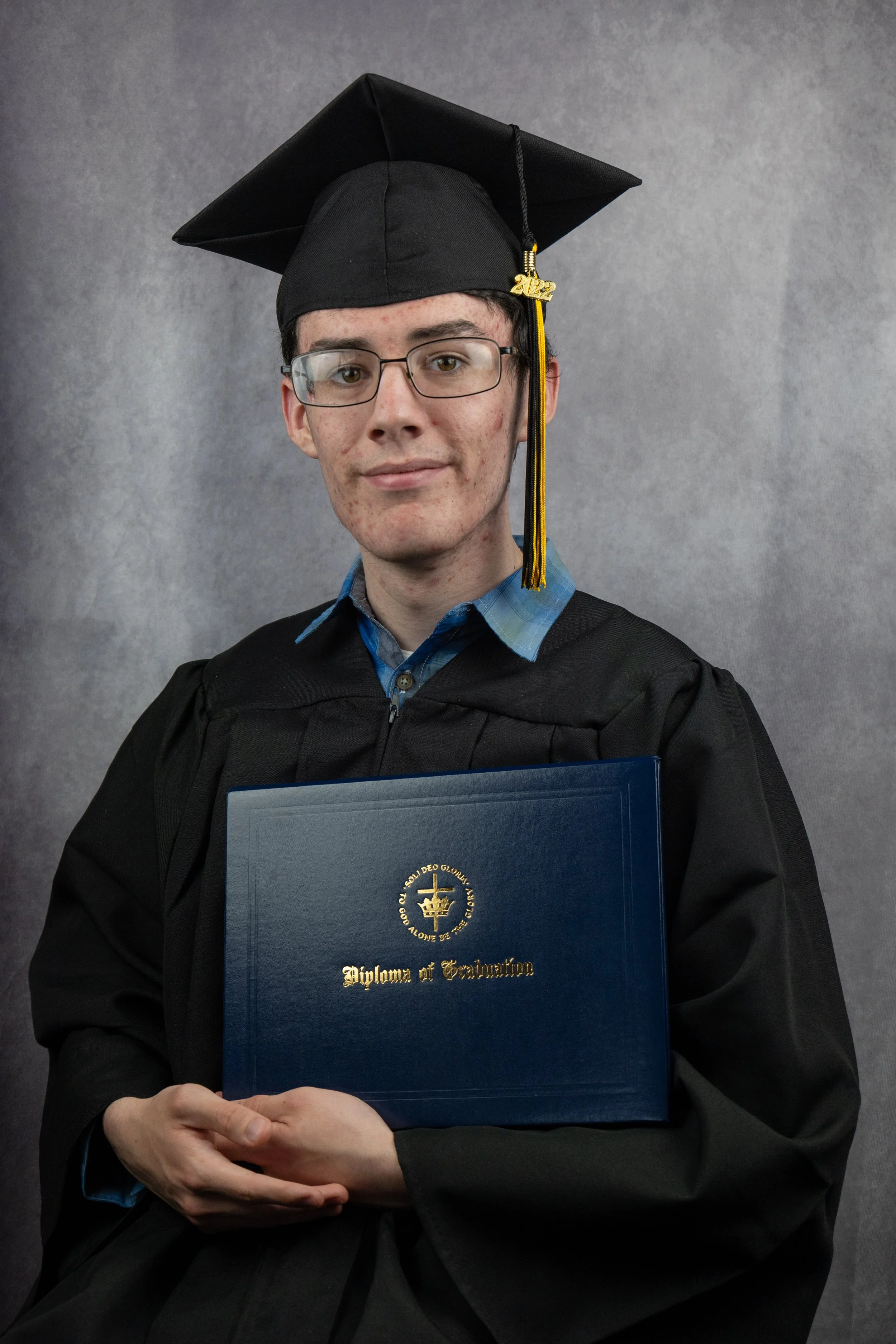 A young man in graduation attire, including a black cap with a gold tassel that says 2022, is holding a diploma in front of him. He is wearing glasses and a blue plaid shirt under his gown. The background is a plain gray.