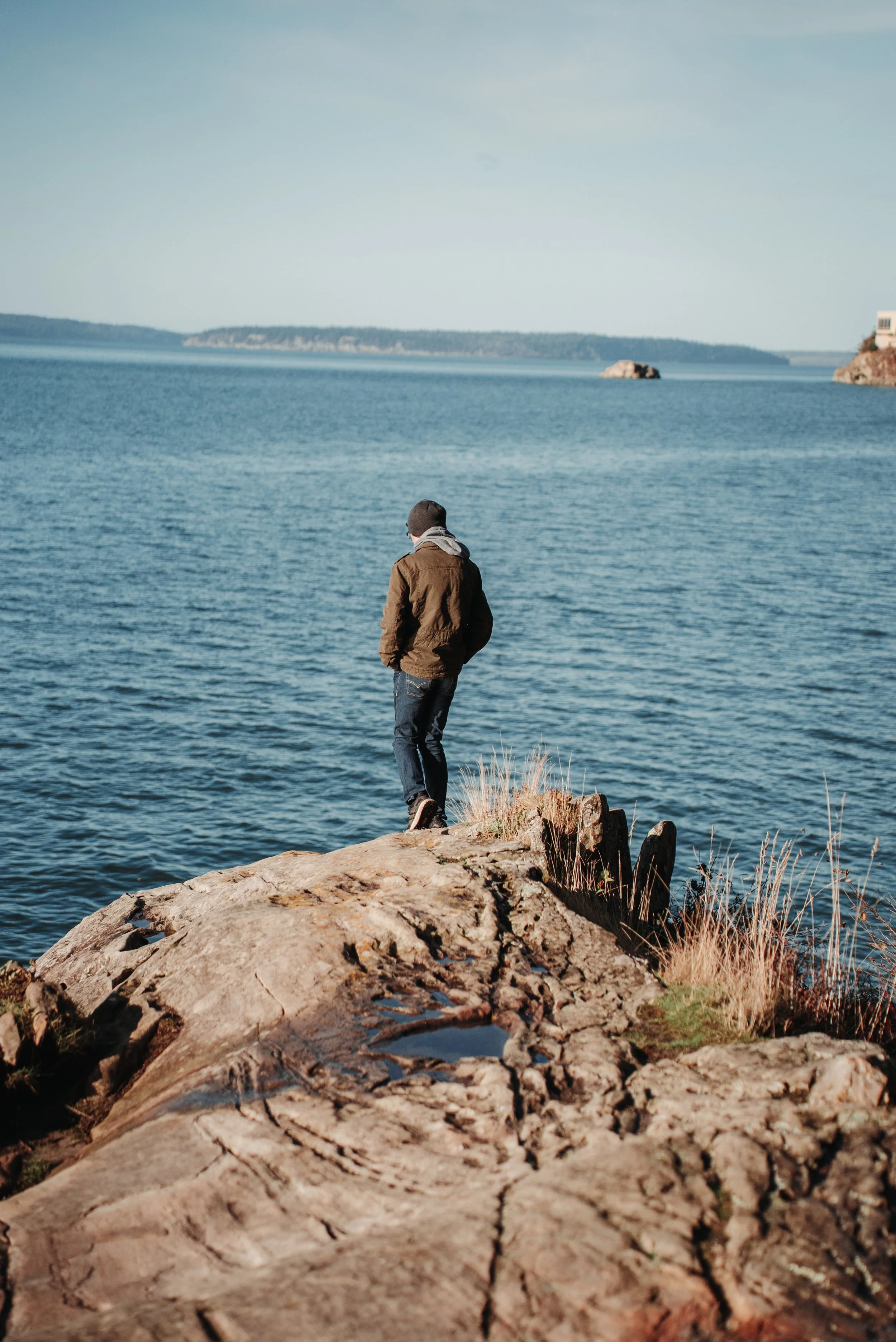A person standing on a rocky shoreline near a body of water, with land in the distance, wearing a brown jacket, black hat, and glasses.