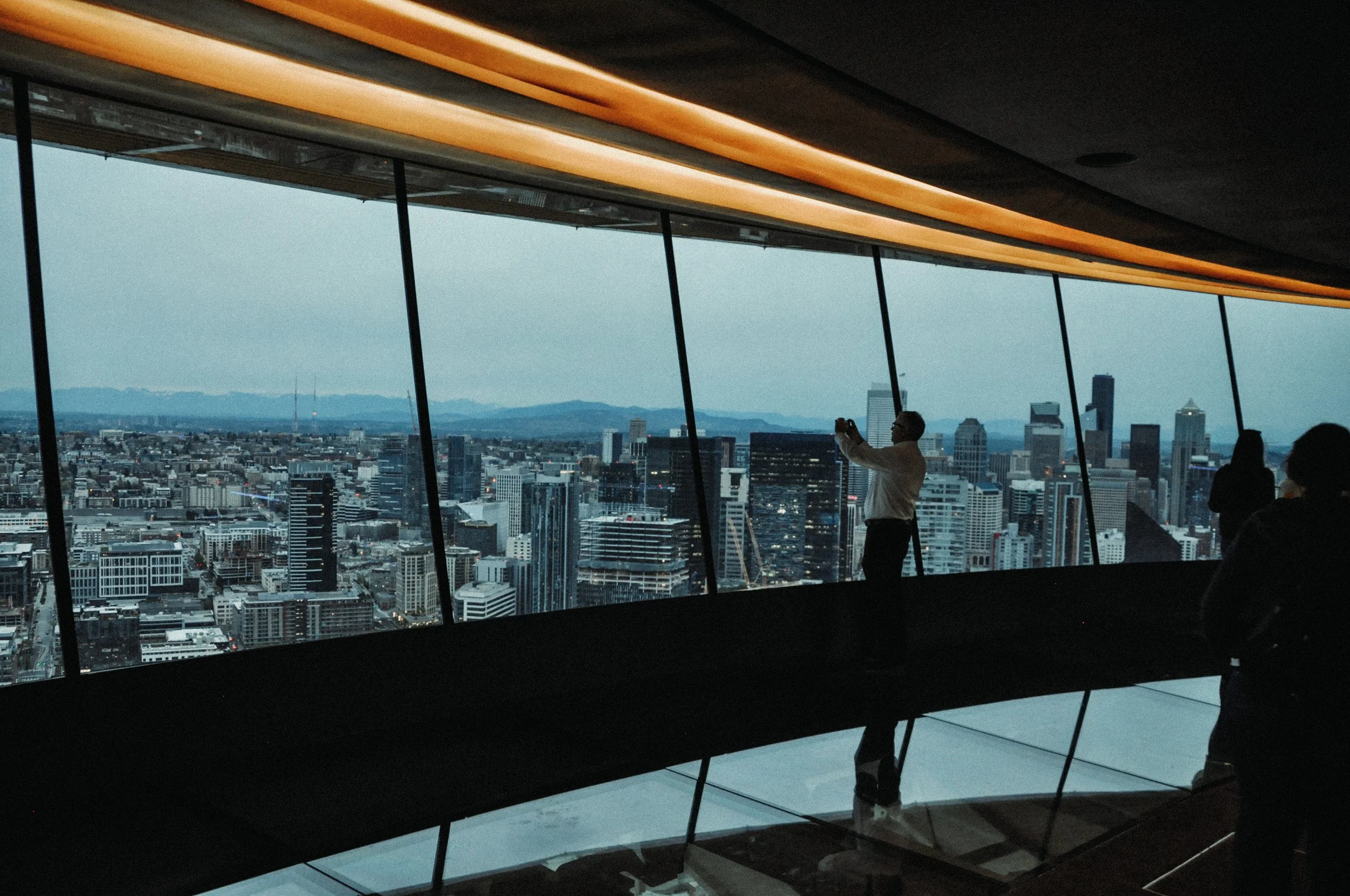 Inside a modern building with large glass windows showing a city skyline, a person taking a photo of the view, and other people visible in the space.