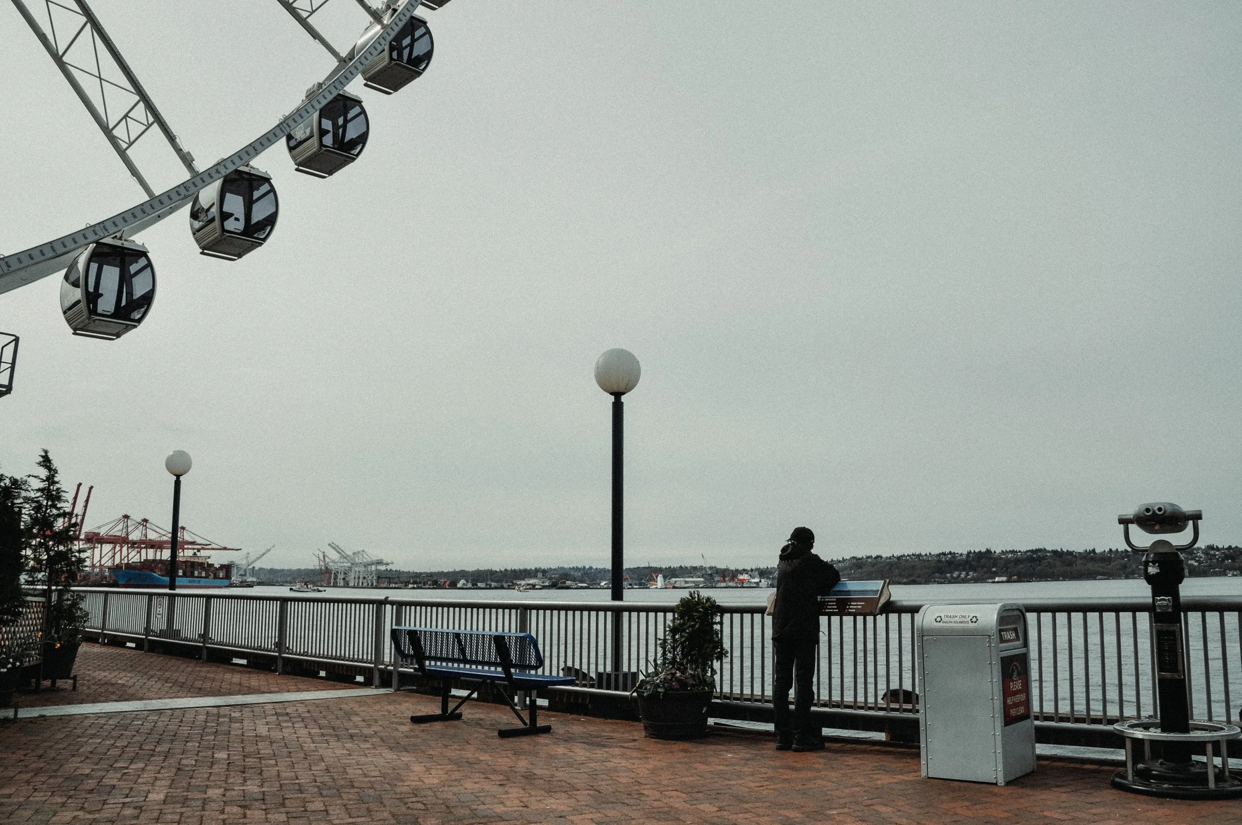 A waterfront scene with a waterfront walkway, benches, street lamps, and a view of ships or boats in the harbor across the water. A person is sitting on a bench, looking at the view. Ferris wheel cabins are visible hanging from the ferris wheel struc
