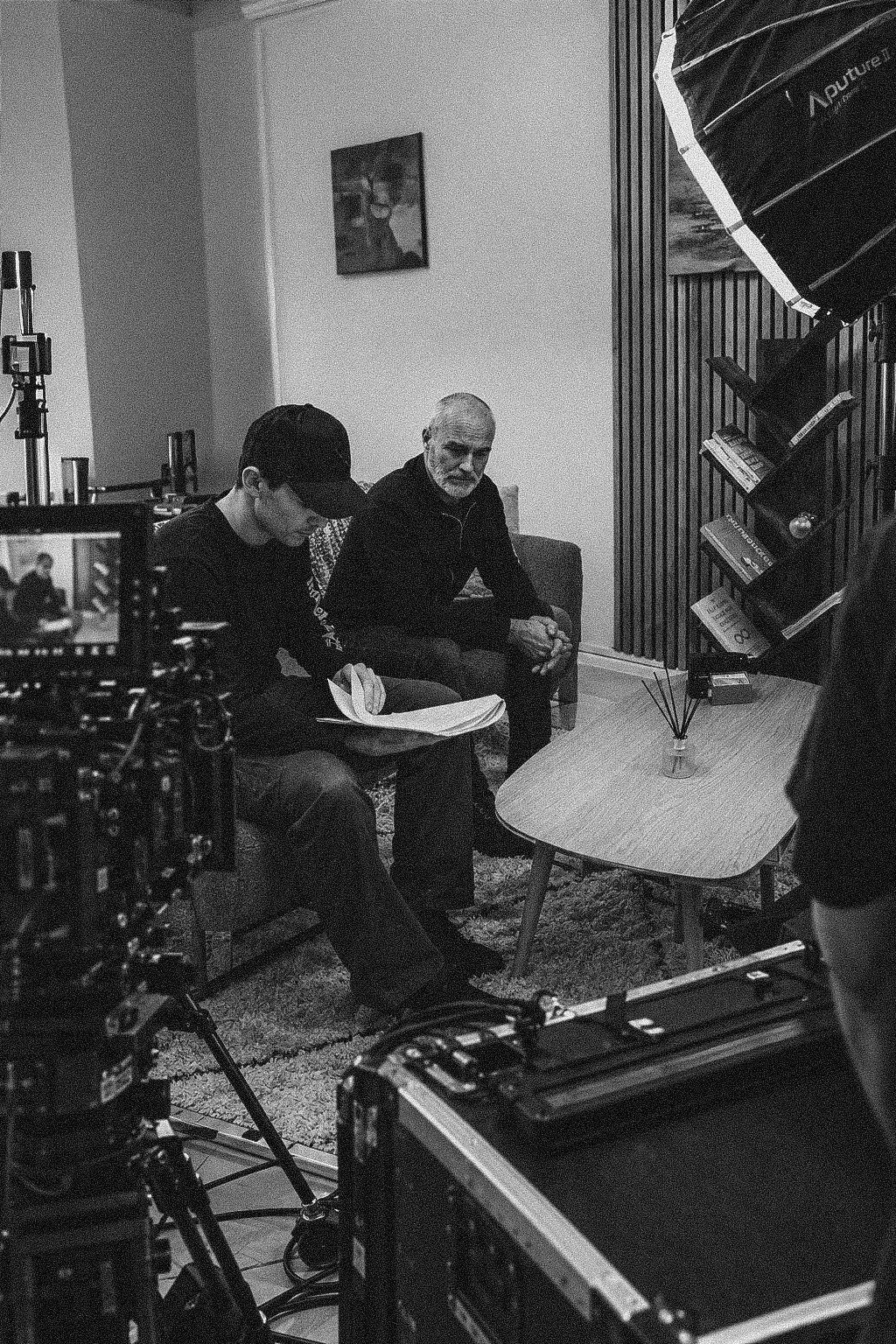 Two men sitting on a sofa in a room with filming equipment visible. One man is reading a script, the other man is looking towards the camera. A camera is recording them, and a softbox light is directed at them. Books and decorative items are on a small coffee table in front of them.