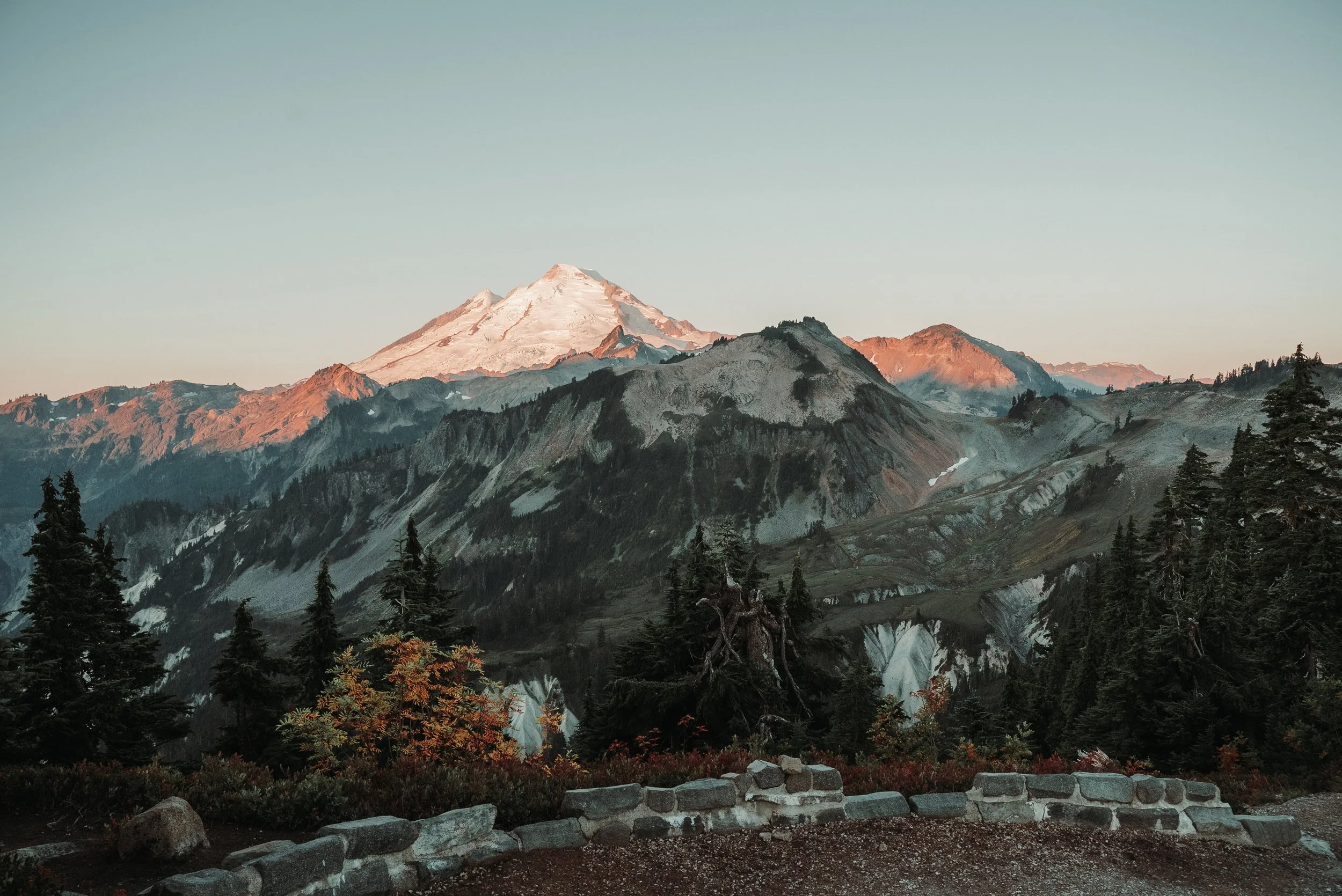Snow-capped mountain peak glowing in the sunset with rocky ridges and forested slopes in the foreground.