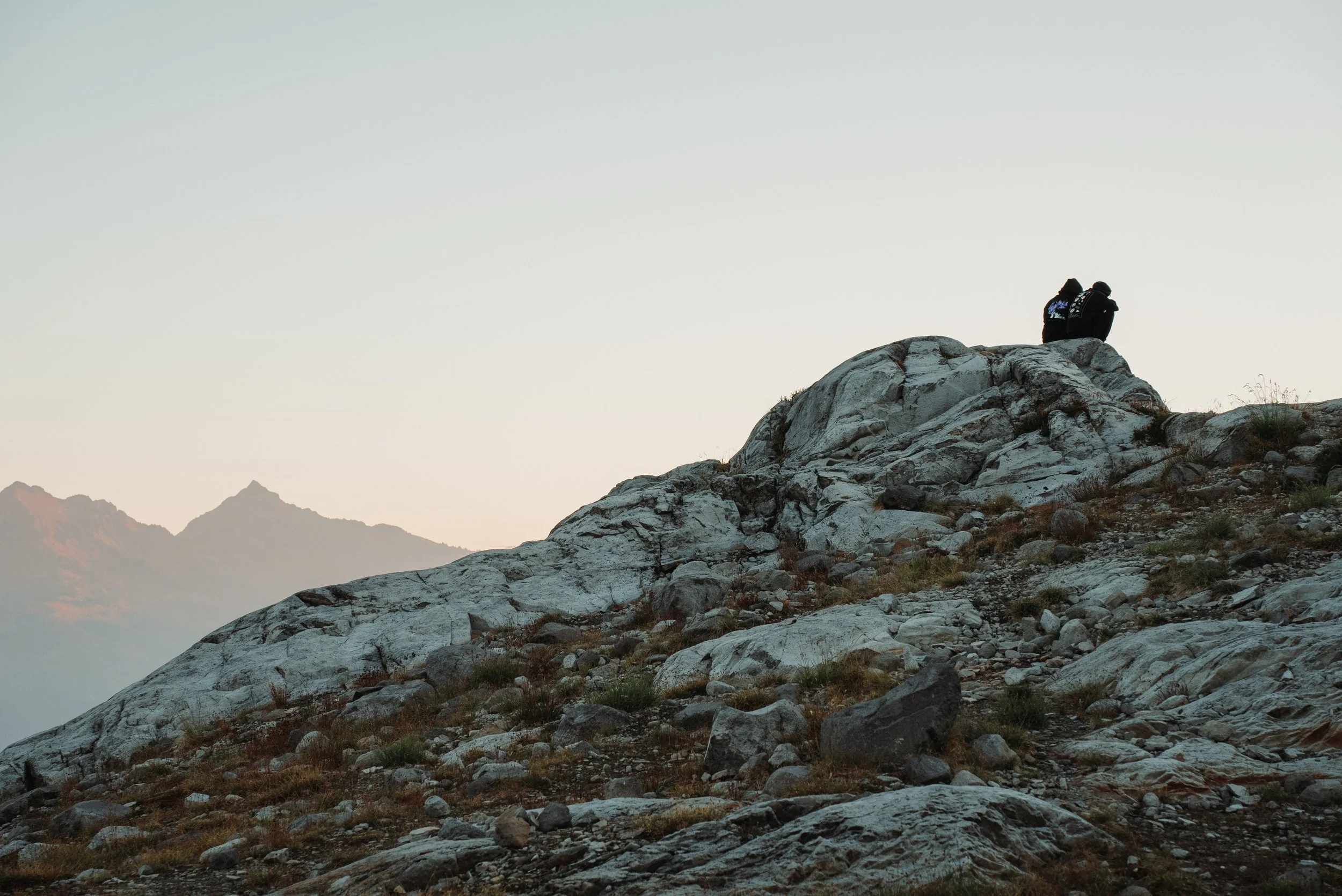 Two people sitting on a rocky mountain top during sunset or sunrise, with mountain peaks in the background.