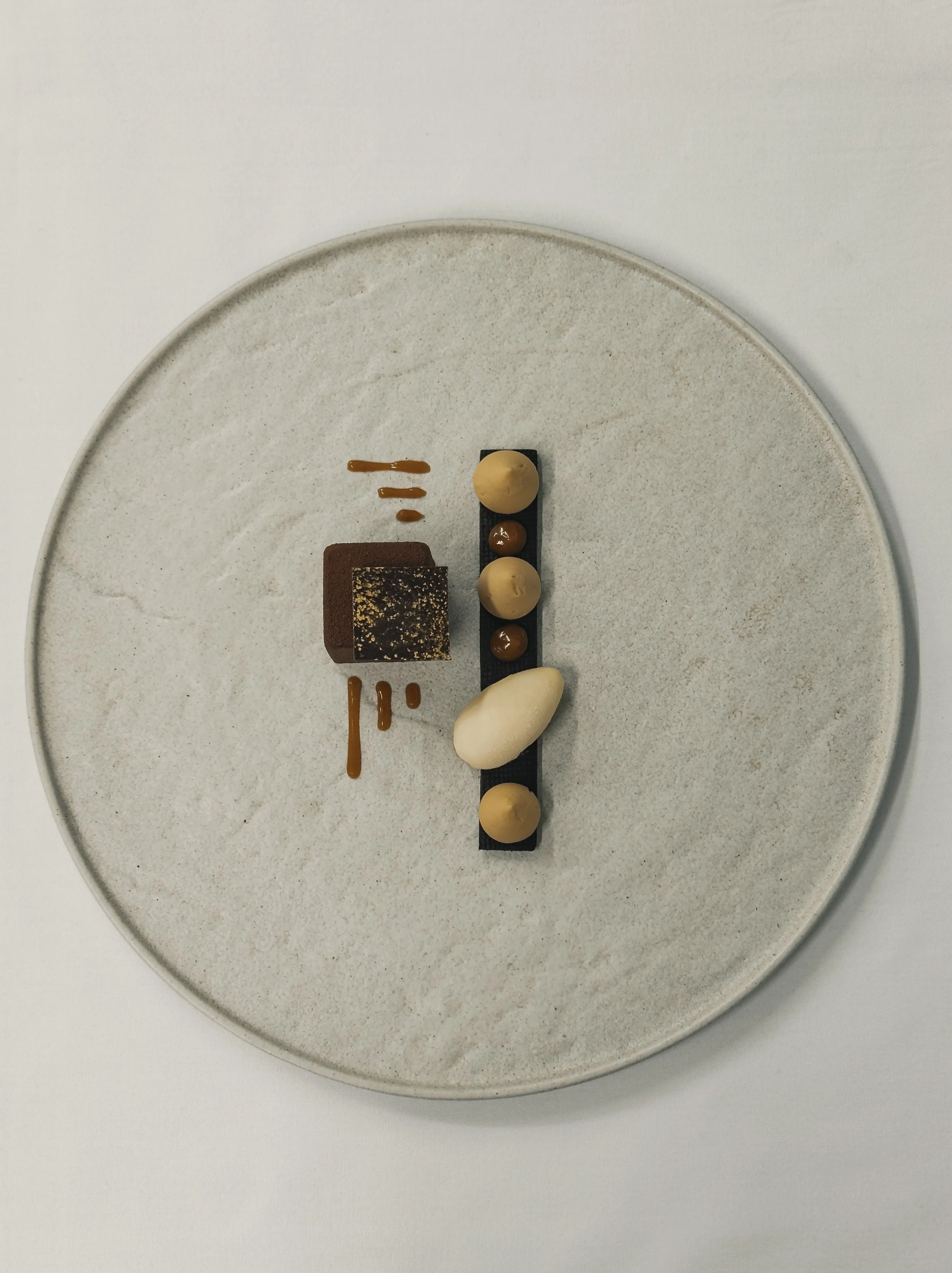 A minimalist dessert plate with a rectangular black chocolate strip, beige and brown dollops, small caramel-like sauces, a square chocolate cake, a white swirl, and caramel drizzle on a white plate.