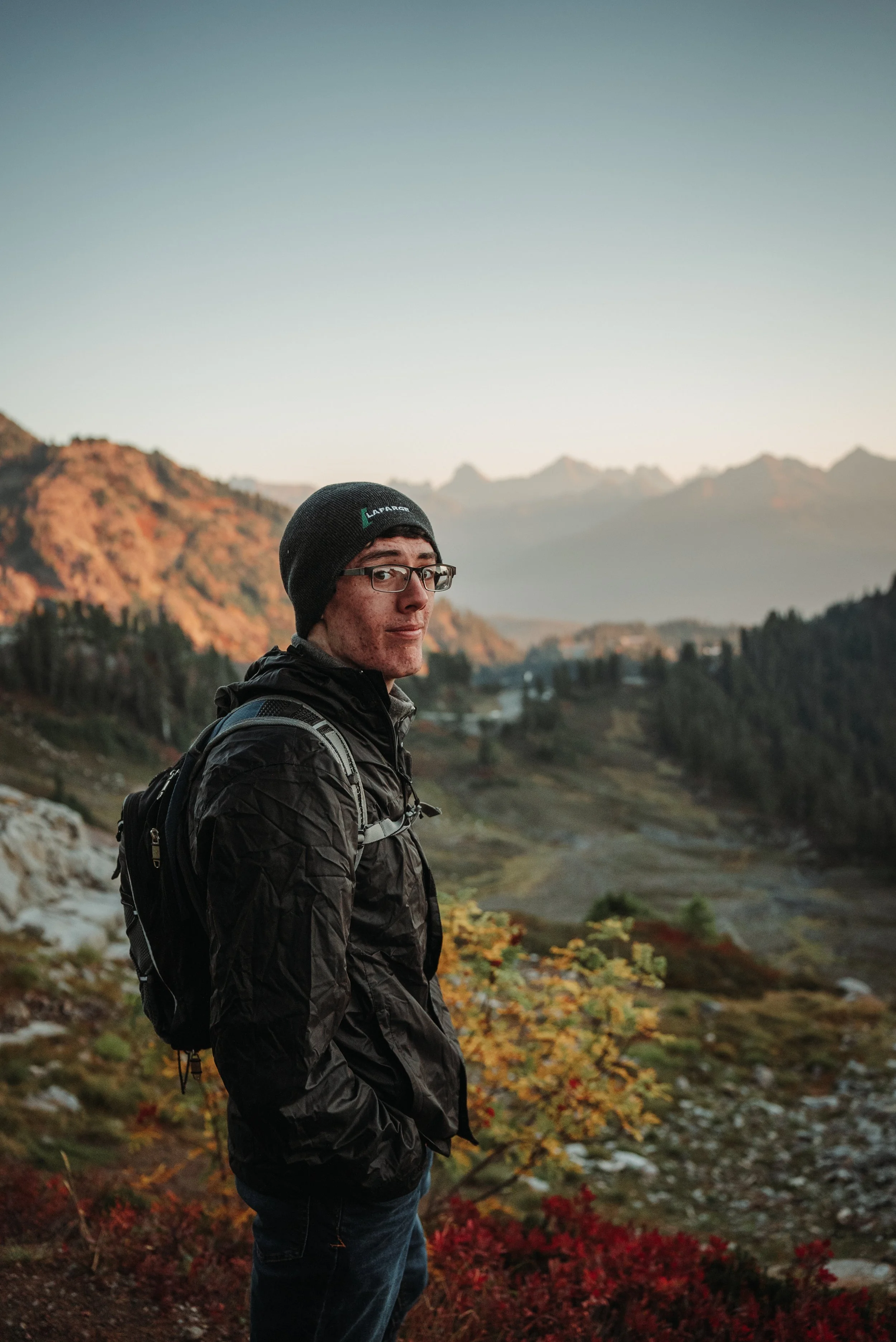 A man wearing a black beanie, glasses, and a black jacket standing outdoors with a mountain landscape behind him during sunset or sunrise.