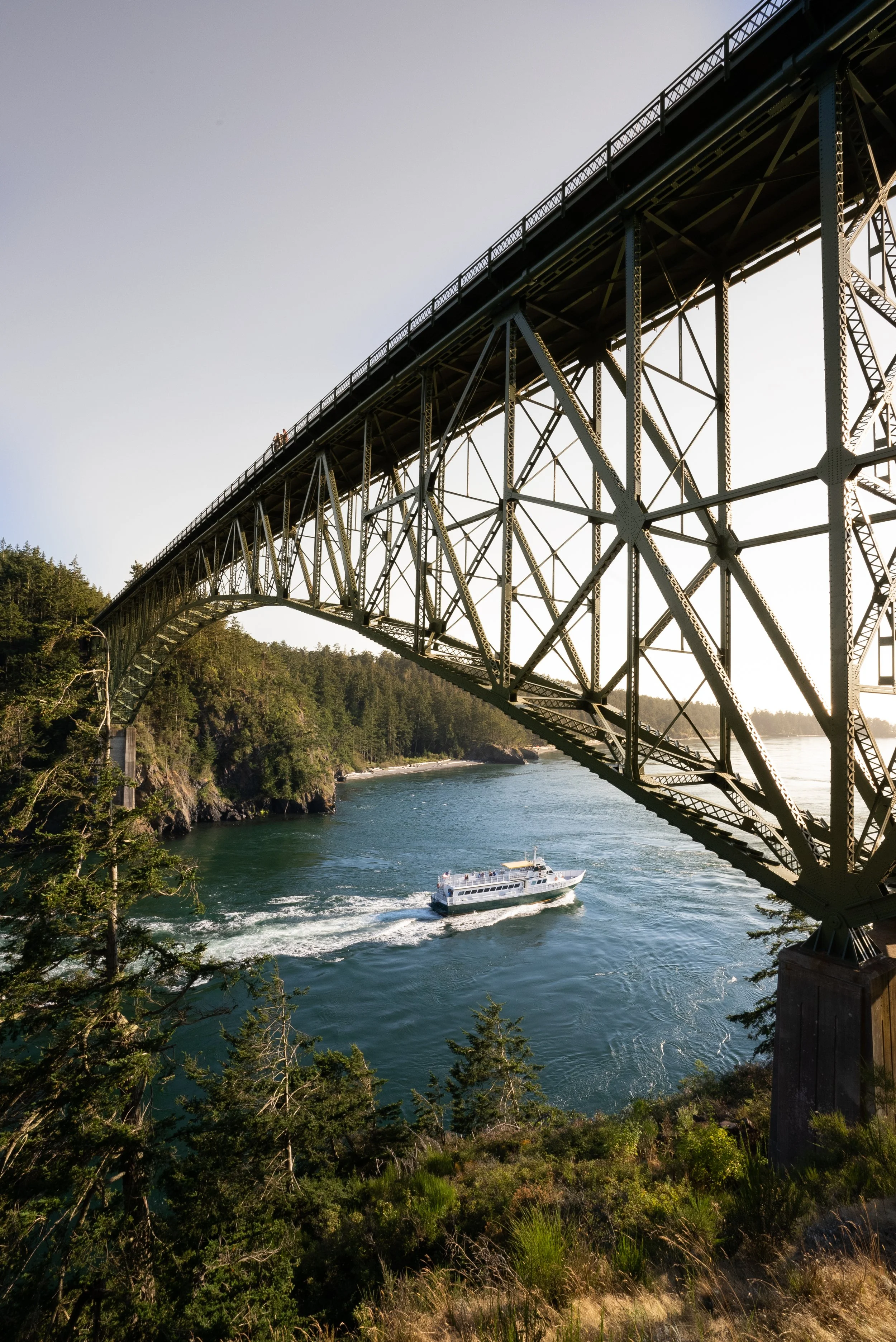A boat sailing under the steel arch of a large bridge over a body of water, with lush green trees and cliffs along the shoreline, and a clear sky in the background.