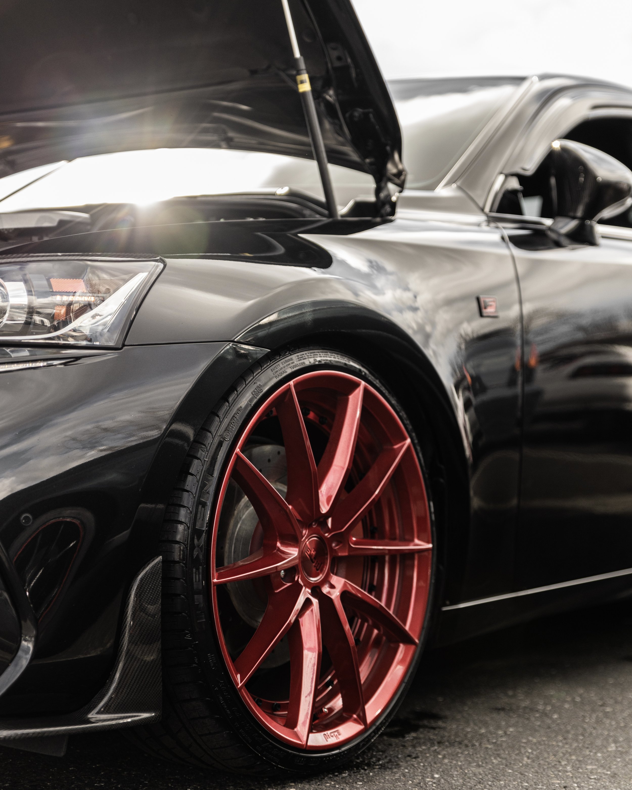Close-up of a black sports car with open hood, showing red custom wheels and a carbon fiber front lip, parked on a dark street.