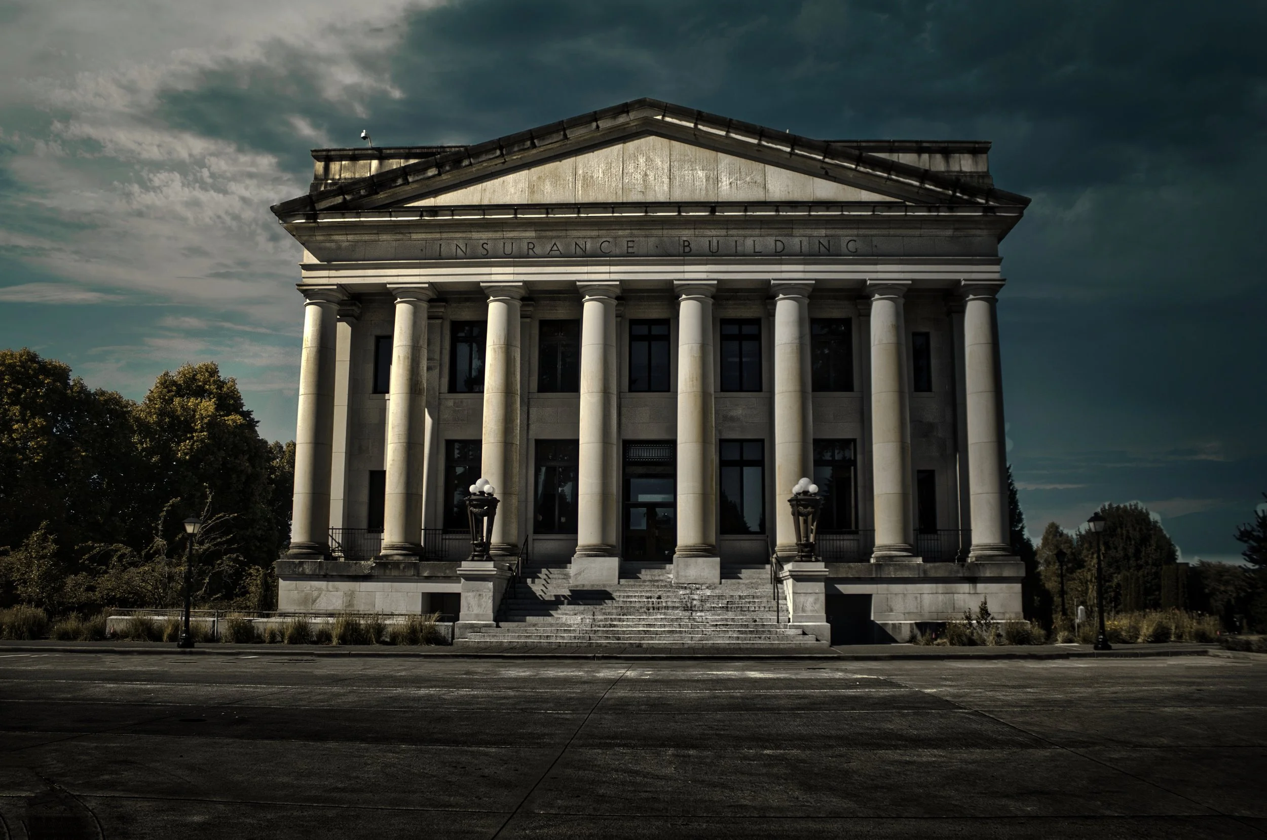 A neoclassical building labeled 'Insurance Building' with tall columns in front, set against a dark, cloudy sky, with a parking area and lampposts in the foreground.