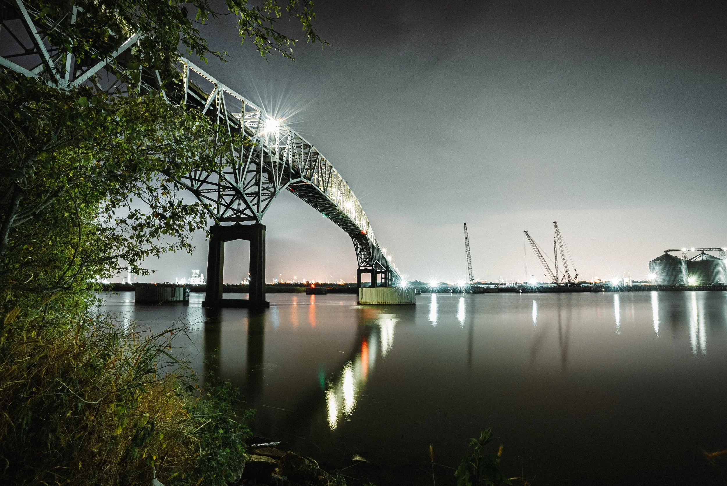 Nighttime view of a large bridge over a body of water, with construction cranes and industrial structures in the background, and trees and vegetation in the foreground.