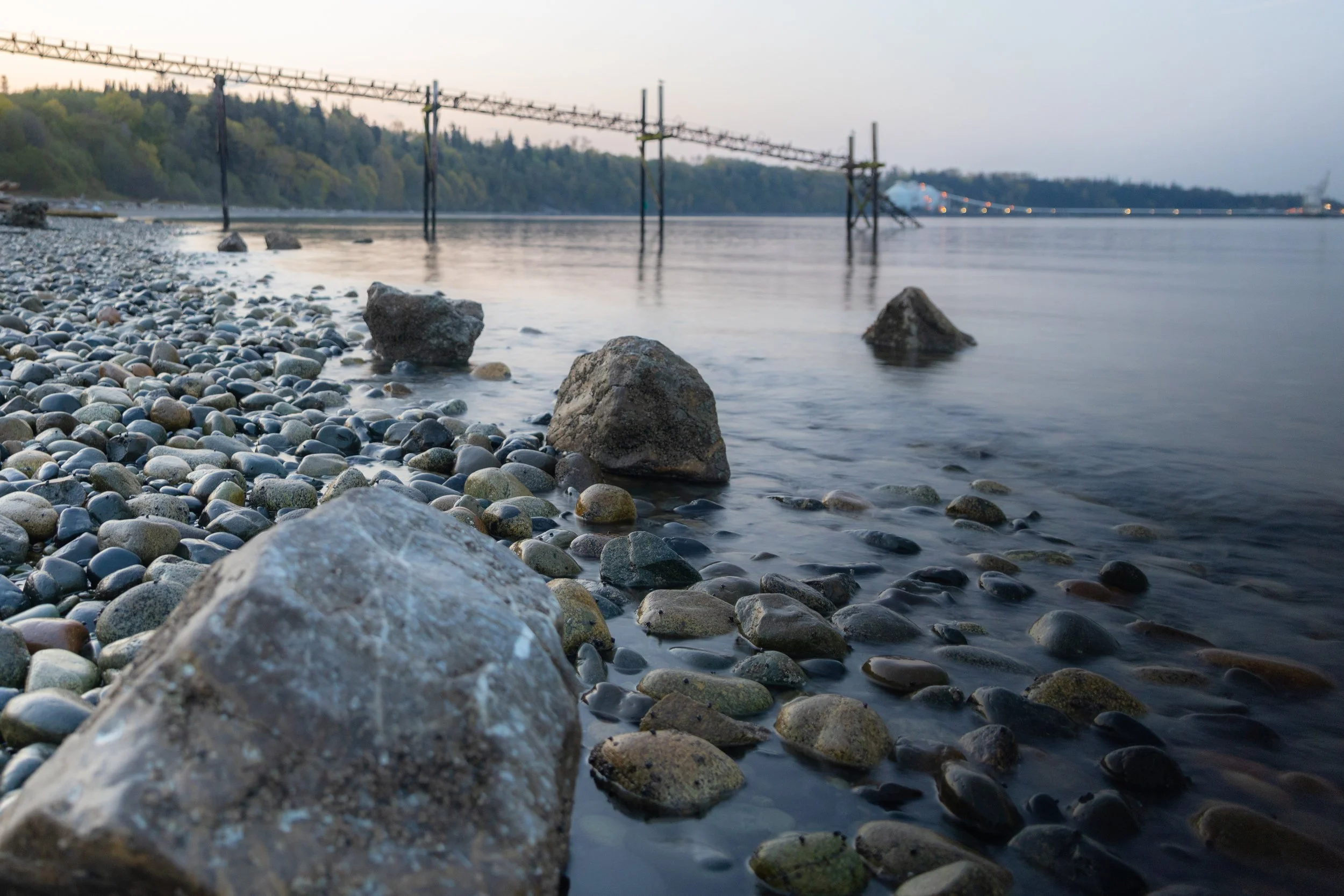 Calm water along a rocky shoreline with large rocks in the foreground and a bridge in the distance, with a forested hillside on the left and foggy sky above.