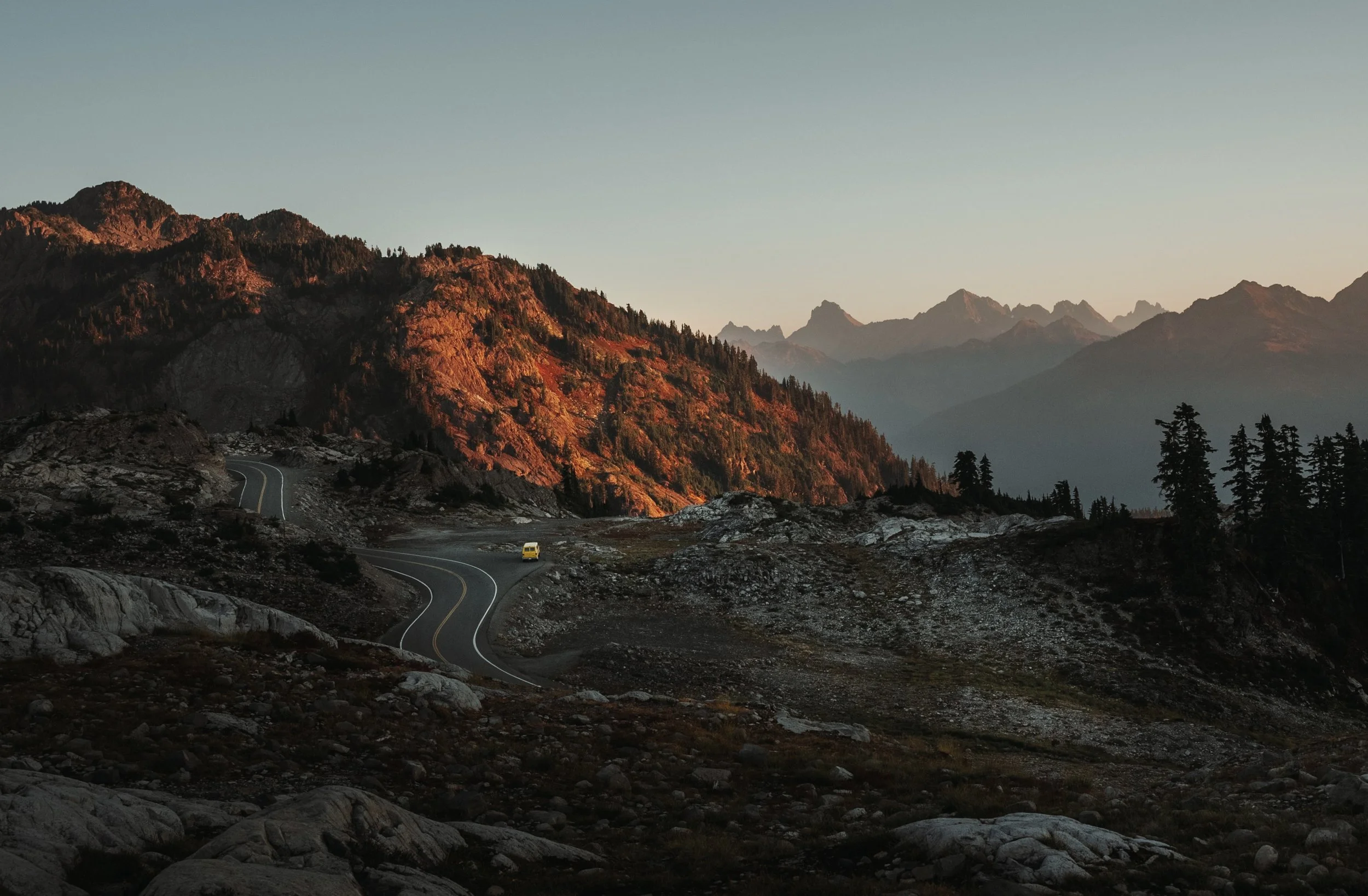 A winding mountain road with a yellow vehicle traveling through rocky terrain, with large mountains covered in trees in the background during sunset or sunrise.