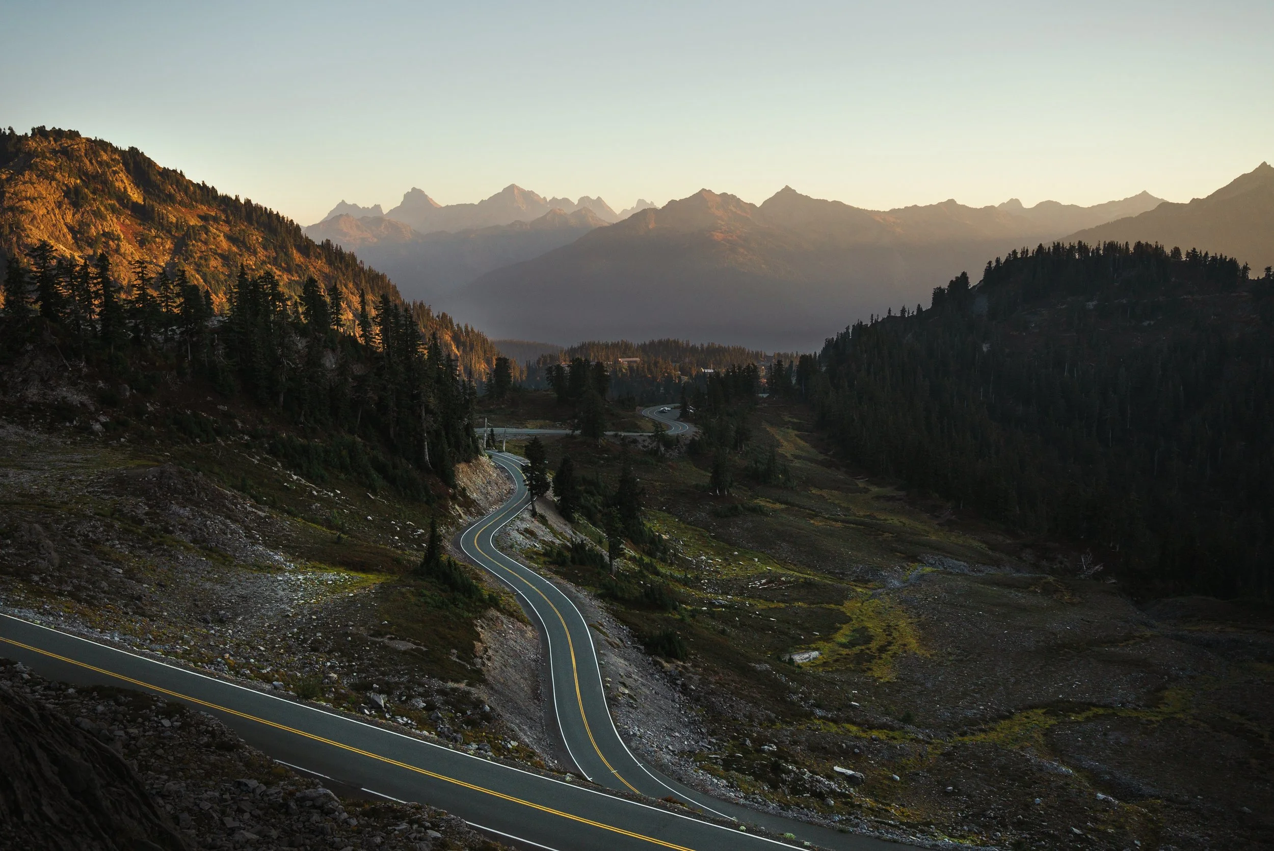 A winding mountain road surrounded by forested hills and distant mountains during sunset.