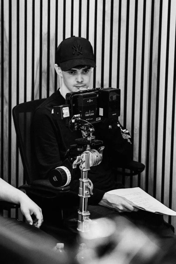 A young man wearing a baseball cap and black shirt sitting in a room with vertical striped walls, operating a camera mounted on a tripod, holding a script or notes.
