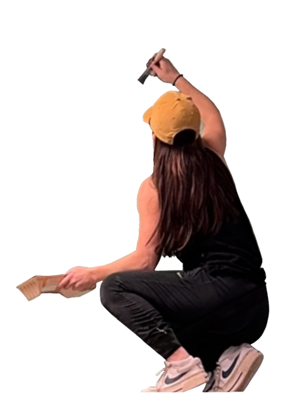 A woman with long brown hair wearing a yellow beanie, black tank top, and black pants, crouching and holding a hammer in one hand and a wooden item in the other.