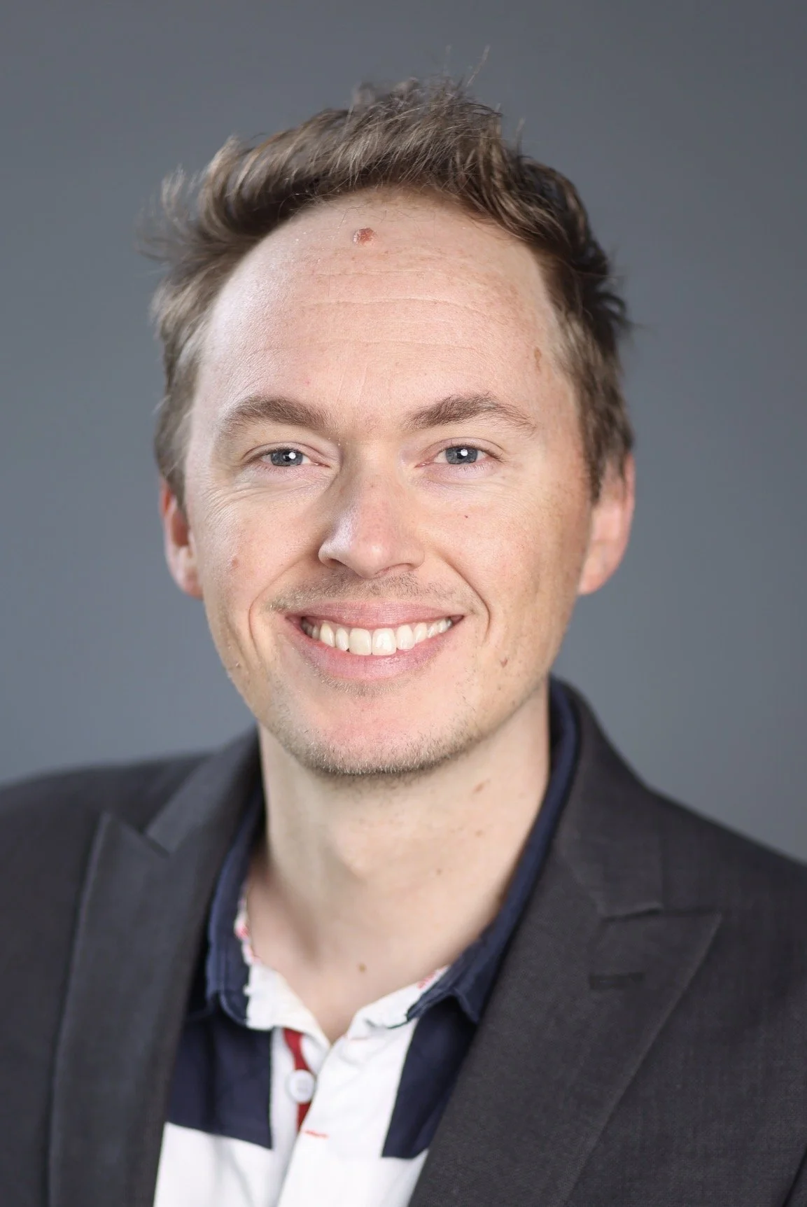 Portrait of a smiling man with short brown hair, light skin, wearing a dark suit and a white shirt with a colorful pattern, against a gray background.
