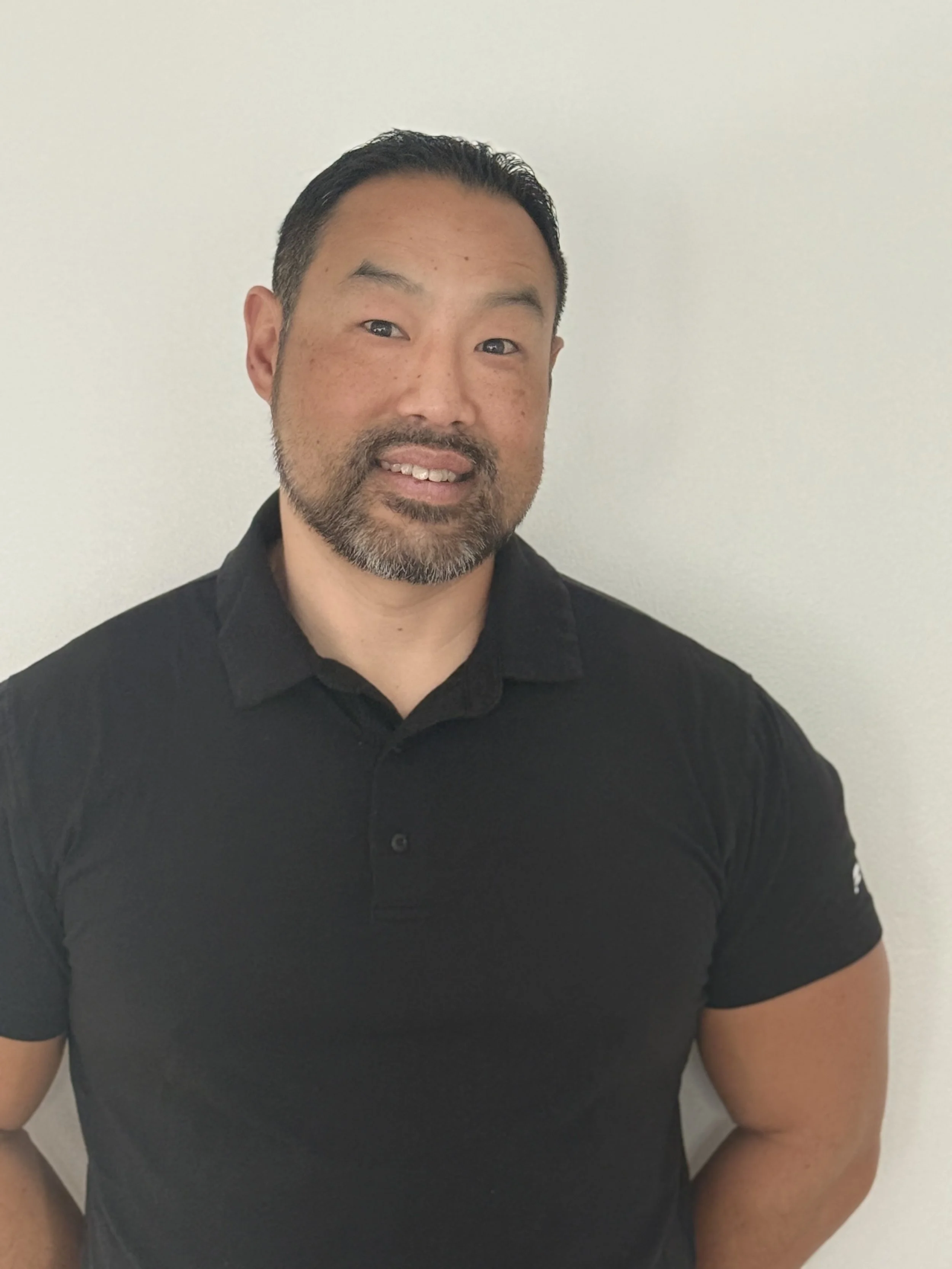 A man with short dark hair and a beard, wearing a black polo shirt, standing against a plain white wall.