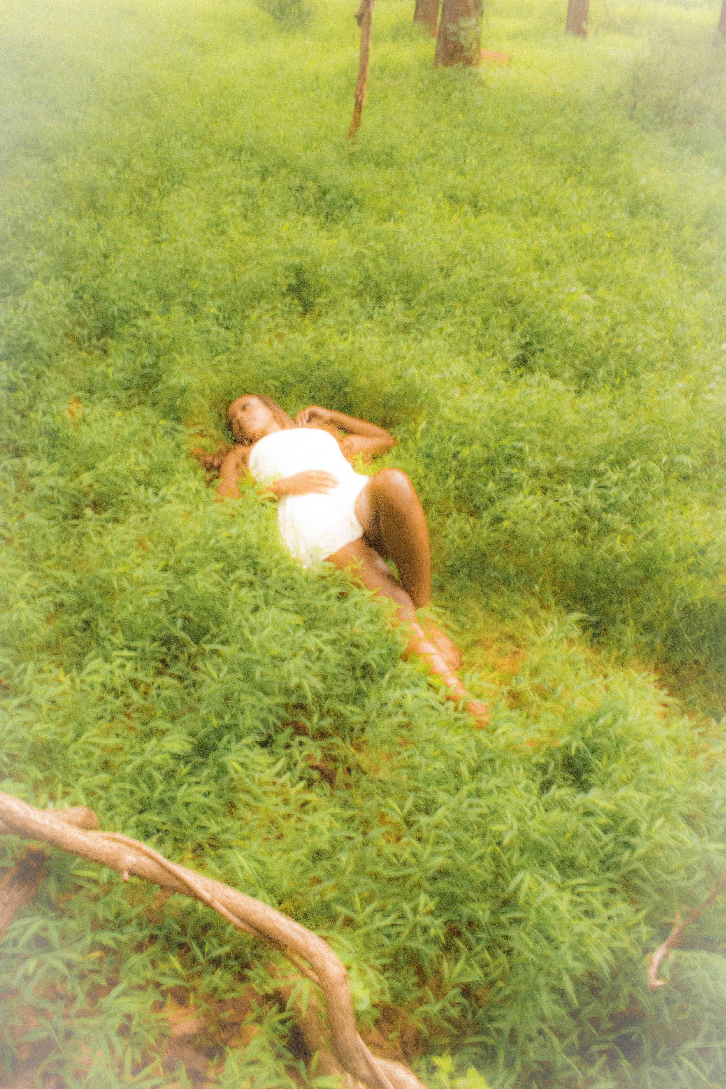 A woman lying on green grass near trees, wearing a white dress.