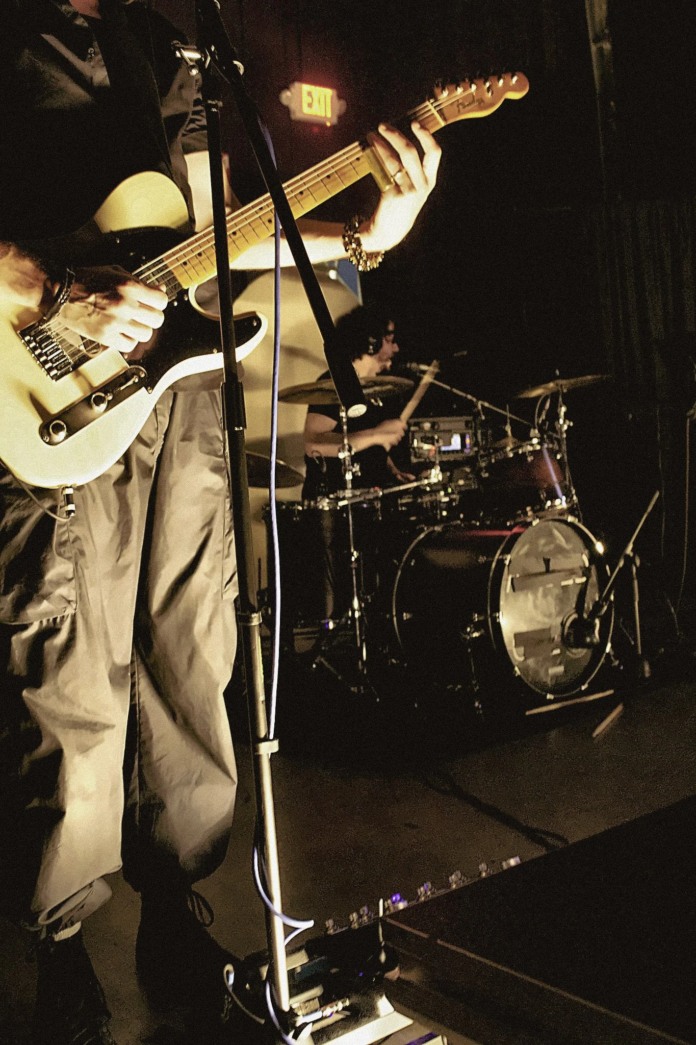 A person playing an electric guitar and a person playing drums in a dimly lit setting, likely a live music performance.