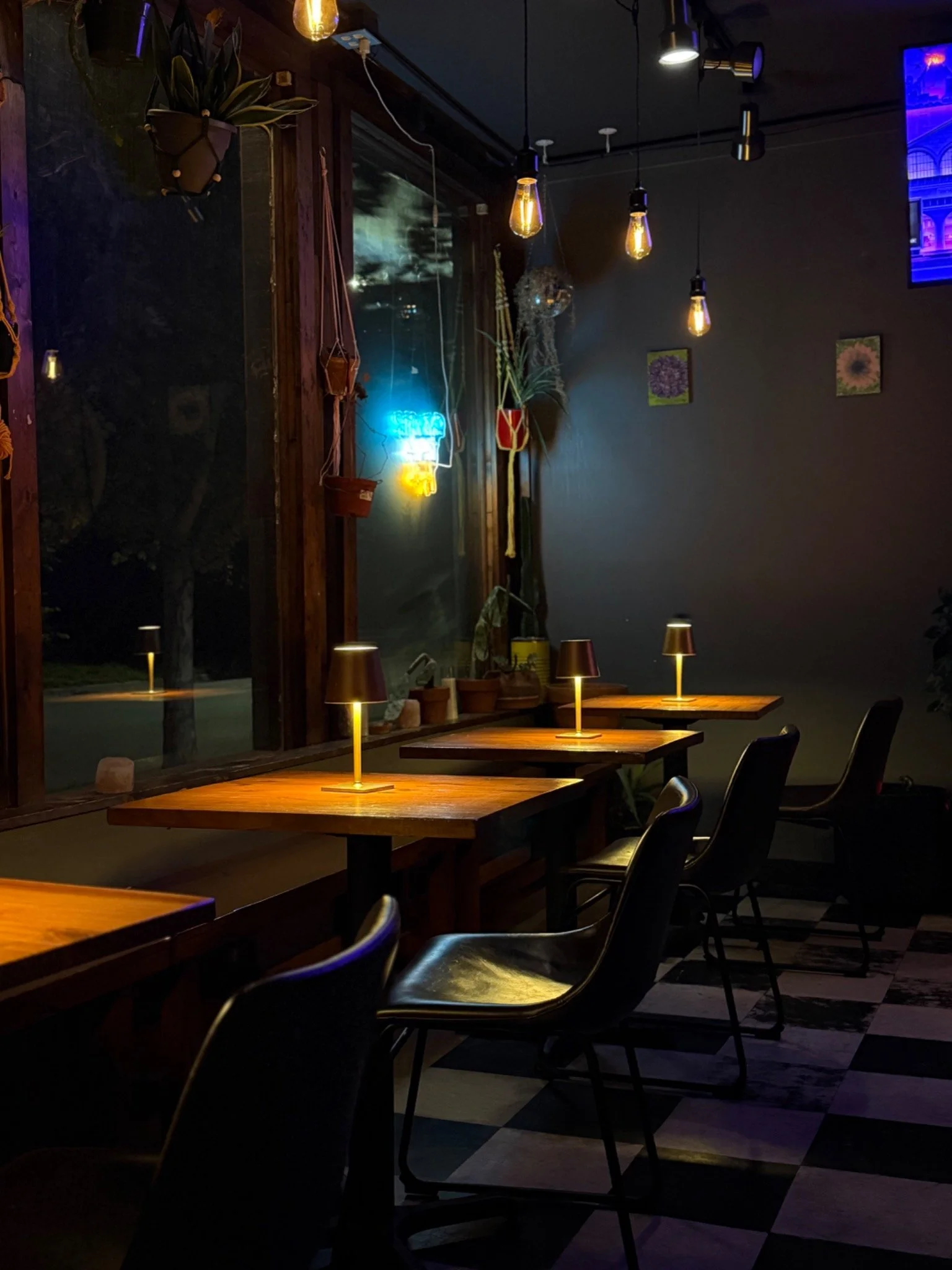 Dimly lit café with wooden tables, black chairs, small lamps on tables, hanging Edison bulbs, a large window with a view of dark outside, and wall decorations including framed pictures of flowers.