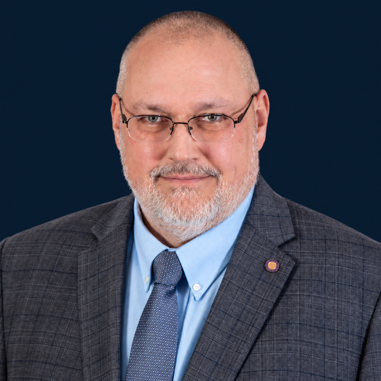 A professional headshot of a middle-aged man with glasses, a beard, and a buzz cut, wearing a dark checked suit, blue shirt, and tie, against a dark blue background.