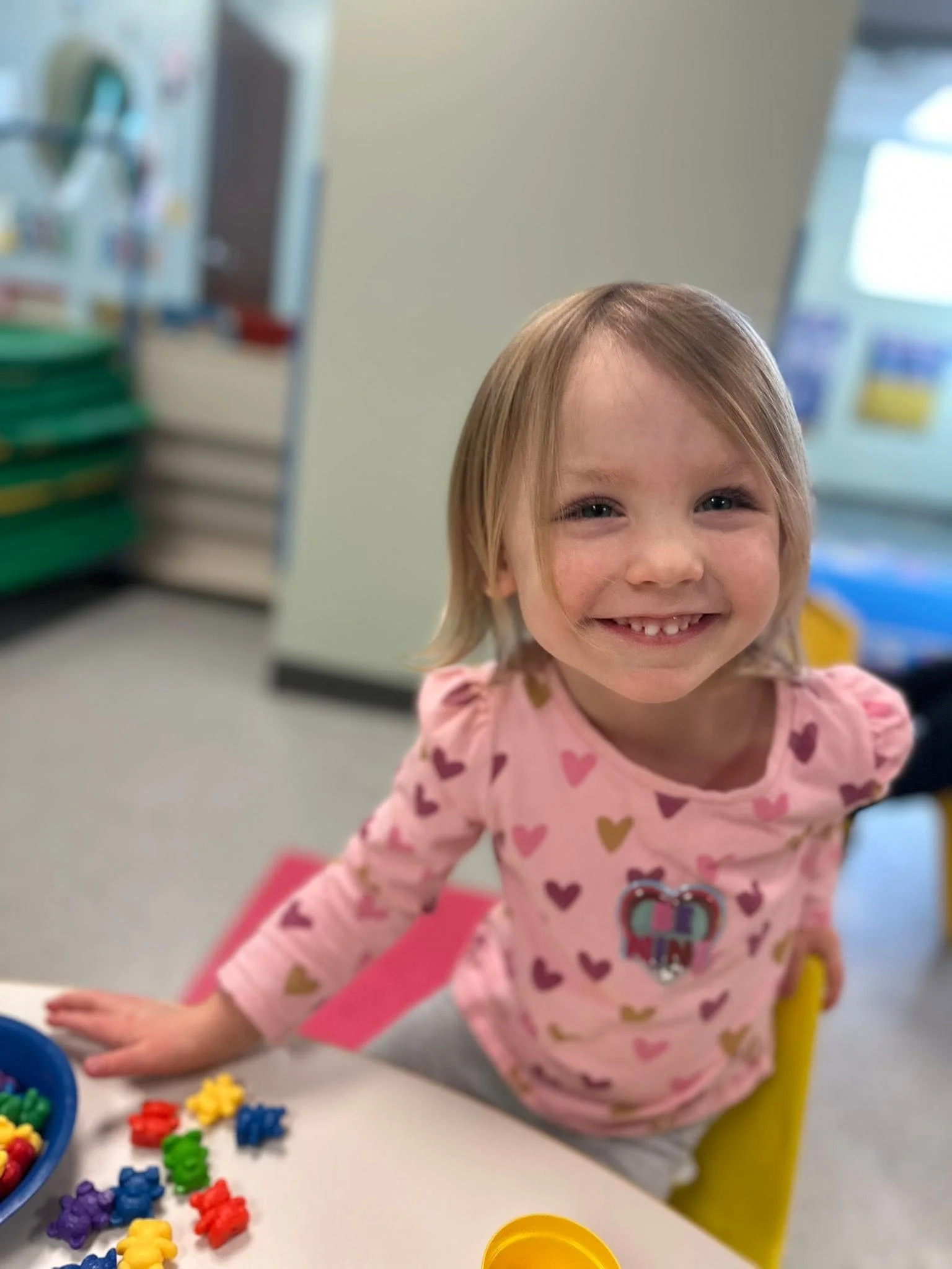 A young girl with blonde hair and a pink shirt with colorful hearts smiling and playing with colorful plastic game pieces on a table in a classroom.
