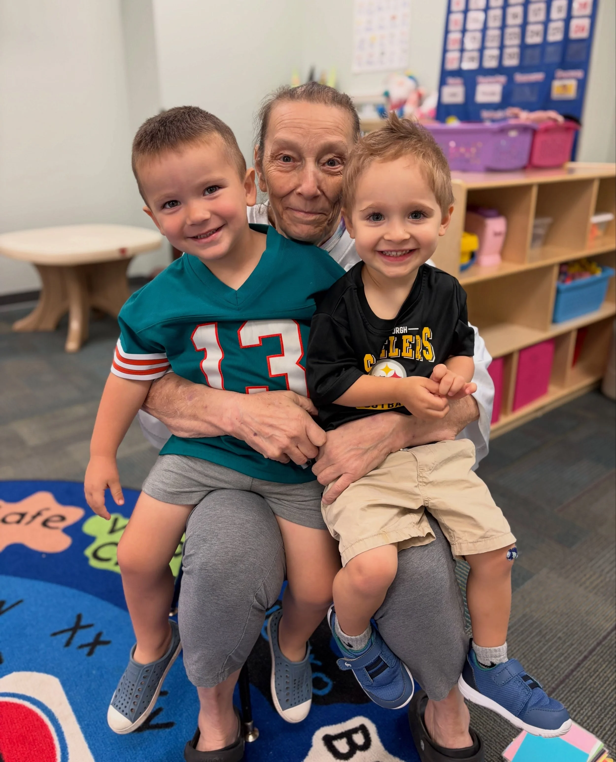 A senior  with short hair and two young boys, one with dark blonde hair and the other with slightly darker blonde hair, smiling and sitting on a woman's lap in a classroom. The background shows colorful posters, a shelf with bins, and educational decor.