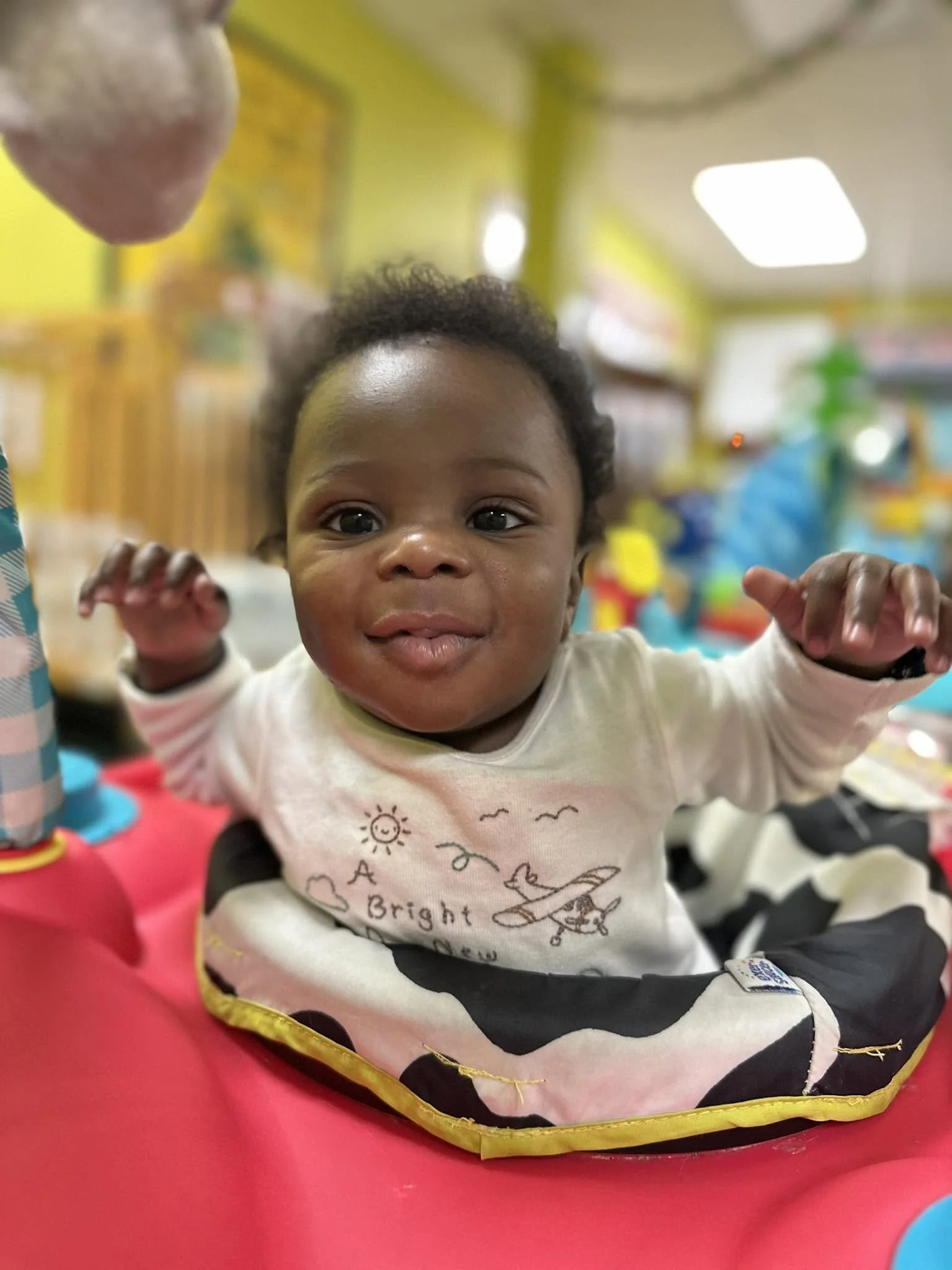 Close-up of a young child's face with a joyful expression, reaching out with hands, in a colorful indoor setting.