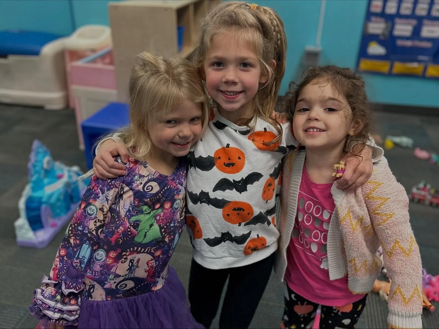 Three young girls standing together, smiling, in a classroom decorated for Halloween, with toys and storage bins in the background.