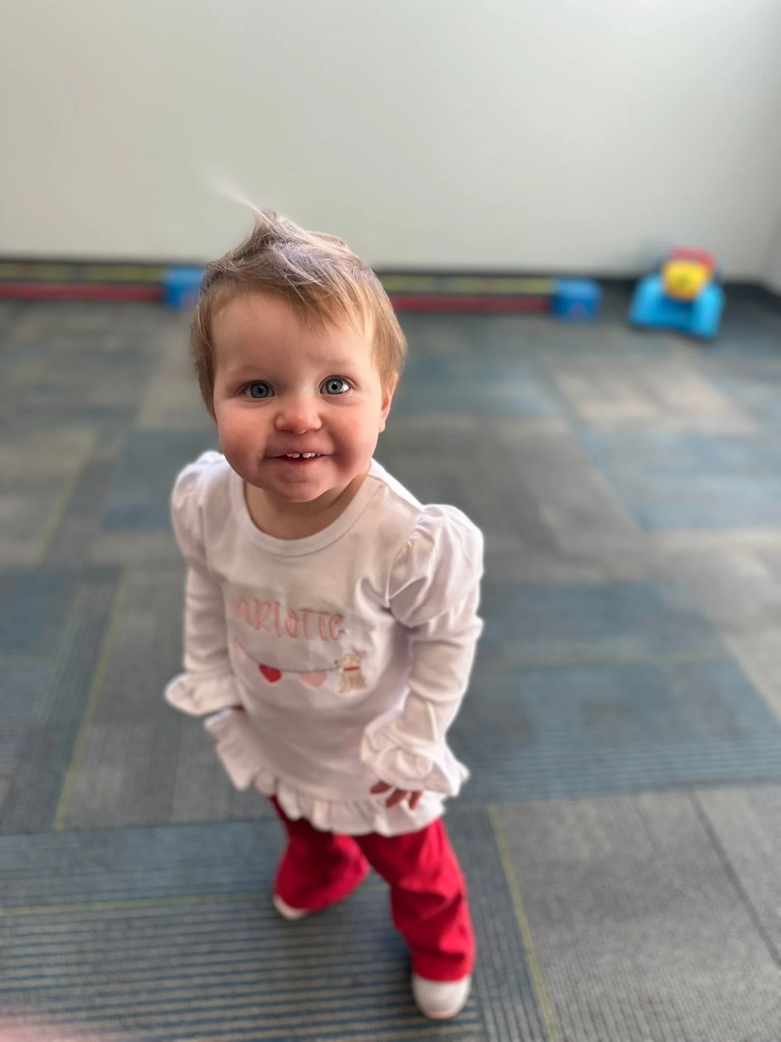 A young child with light brown hair, blue eyes, and a big smile, is standing indoors on a blue carpet. The child is wearing a white shirt with red and pink letters, red pants, and white shoes. In the background, there are colorful toys and a white wall.