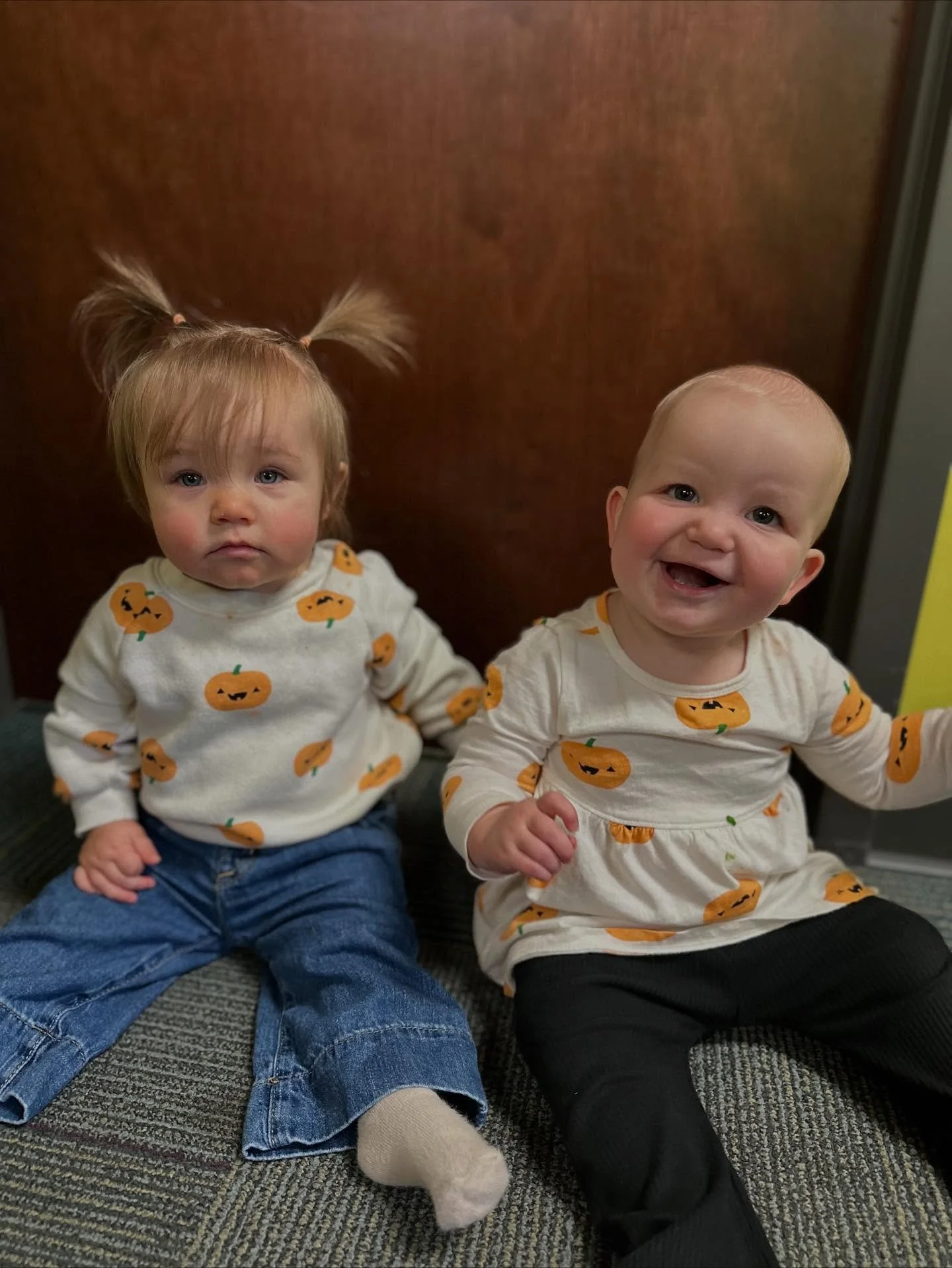 Two young children sitting on the floor in front of a wooden door, wearing matching Halloween pumpkin-themed shirts. The girl on the left has light brown hair styled in pigtails and a serious expression. The  on the right has blonde hair and is smiling.