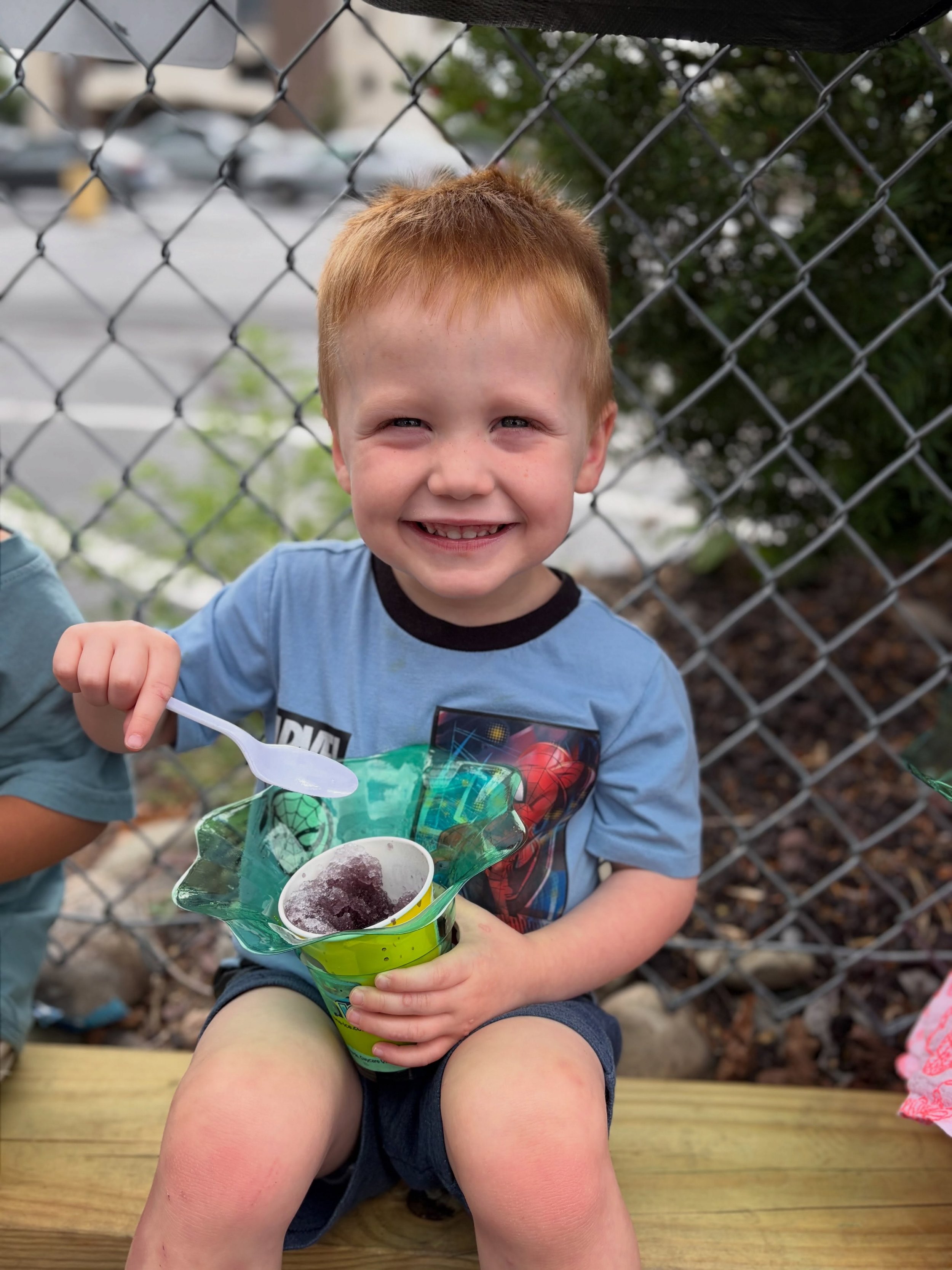 Smiling young boy with red hair sitting outdoors near a chain link fence, holding a colorful ice cream cone container, wearing a blue Marvel T-shirt, with a wooden platform and blurred background of trees and cars.