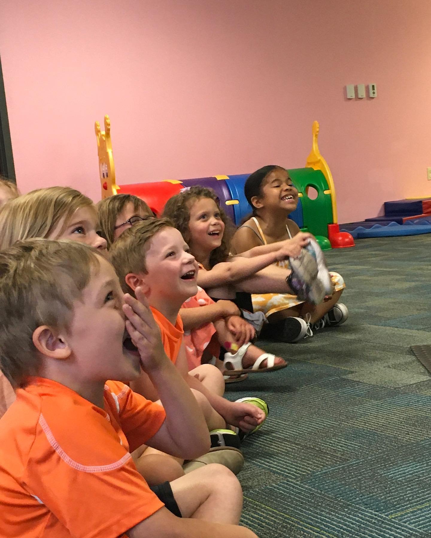 A group of children sitting on the floor, looking and smiling excitedly at something off-camera in a room with pink walls and colorful play equipment.