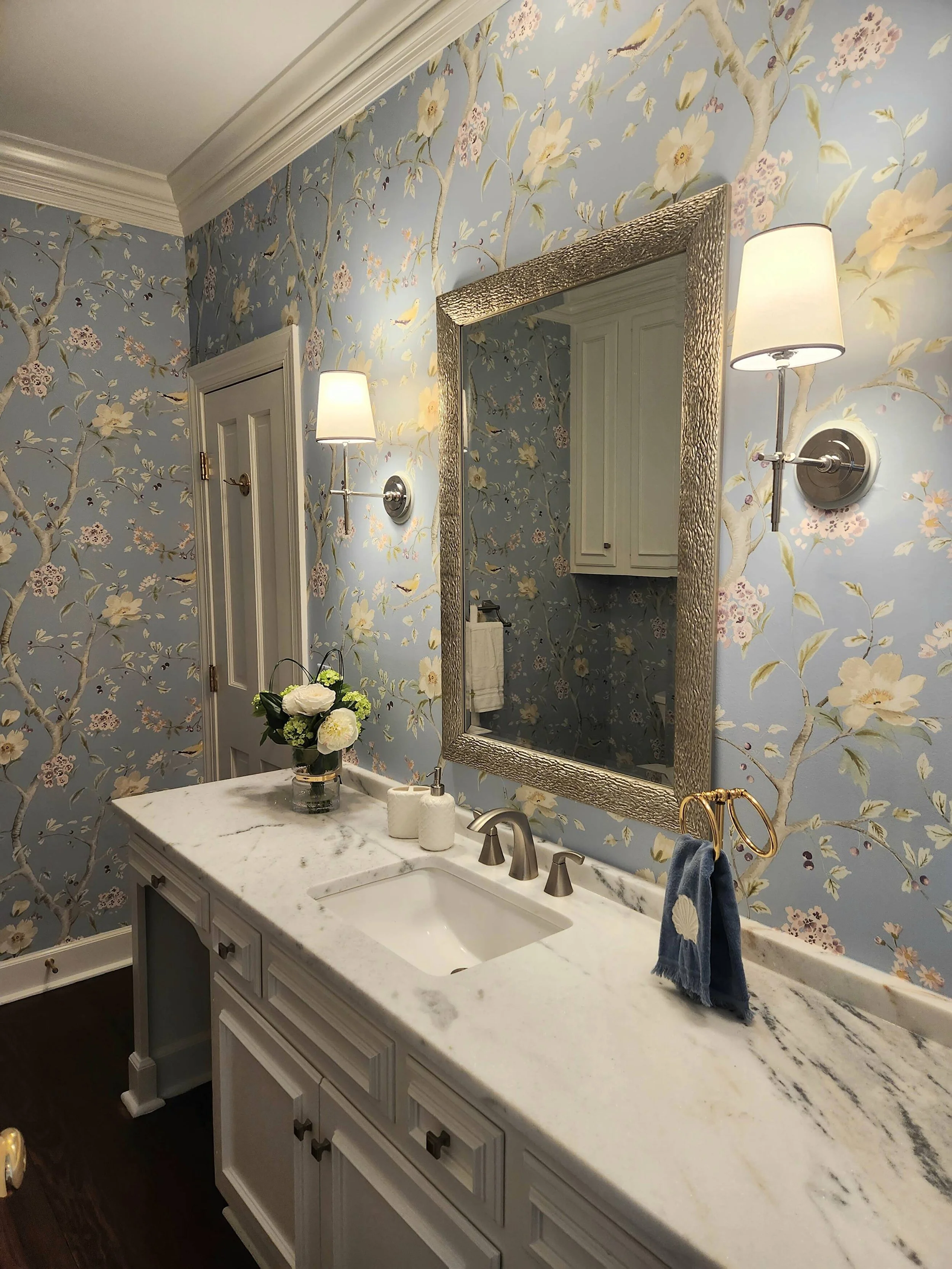 Bathroom vanity with blue patterned wallpaper, a gilded mirror, and brass sconces designed by Bonnie Boyette Designs