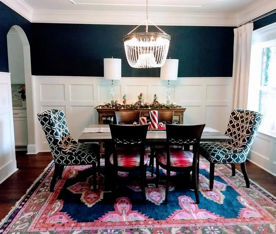 Dining room with navy walls, white wainscoting, patterned host chairs, and a vibrant rug designed by Bonnie Boyette Designs