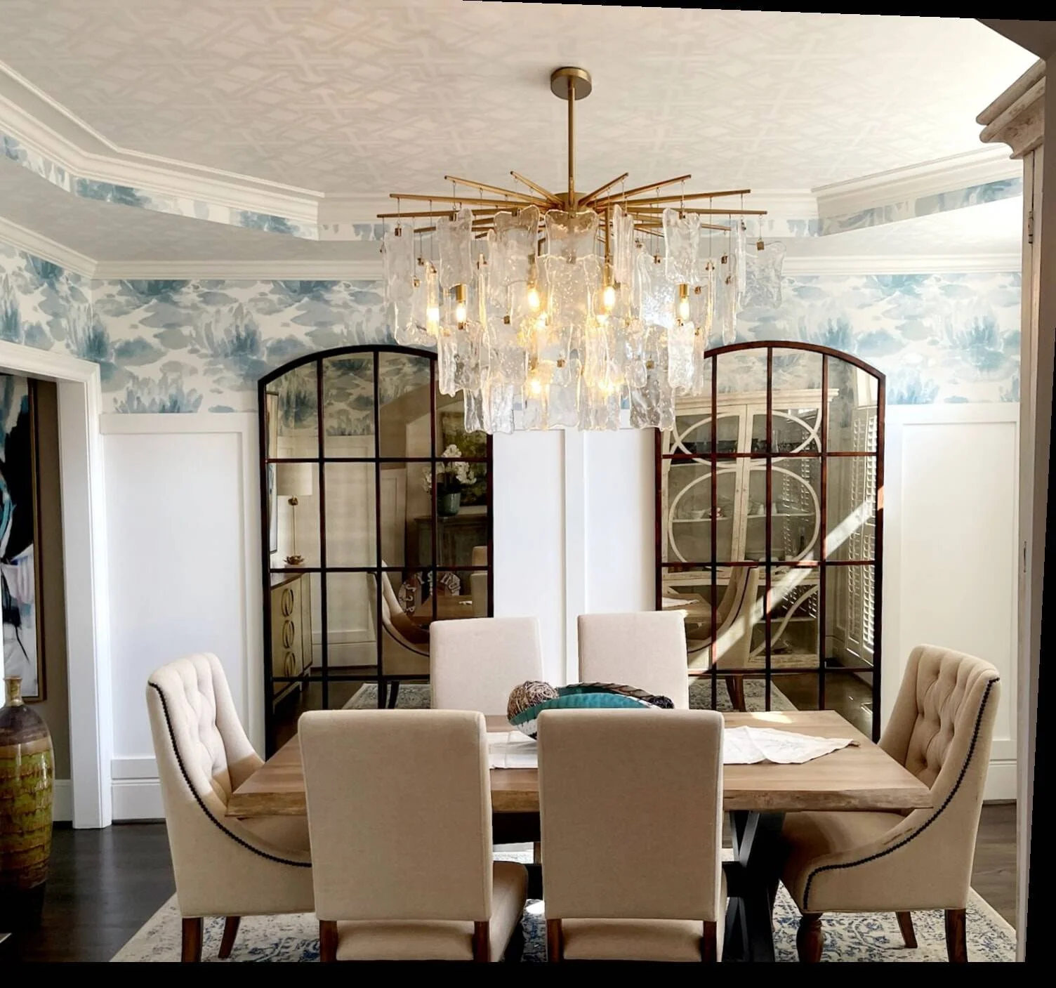 Formal dining room with a glass chandelier, arched black mirrors, blue toile wallpaper, and tufted chairs designed by Bonnie Boyette Designs