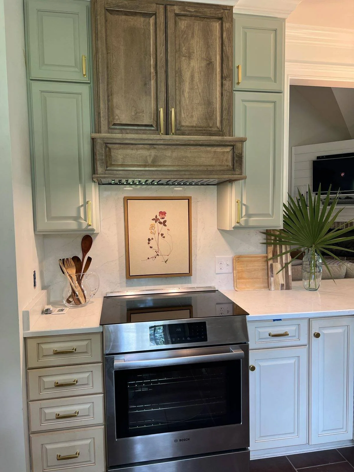 Kitchen range wall with a wood hood, sage green cabinets, and framed art on the backsplash designed by Bonnie Boyette Designs