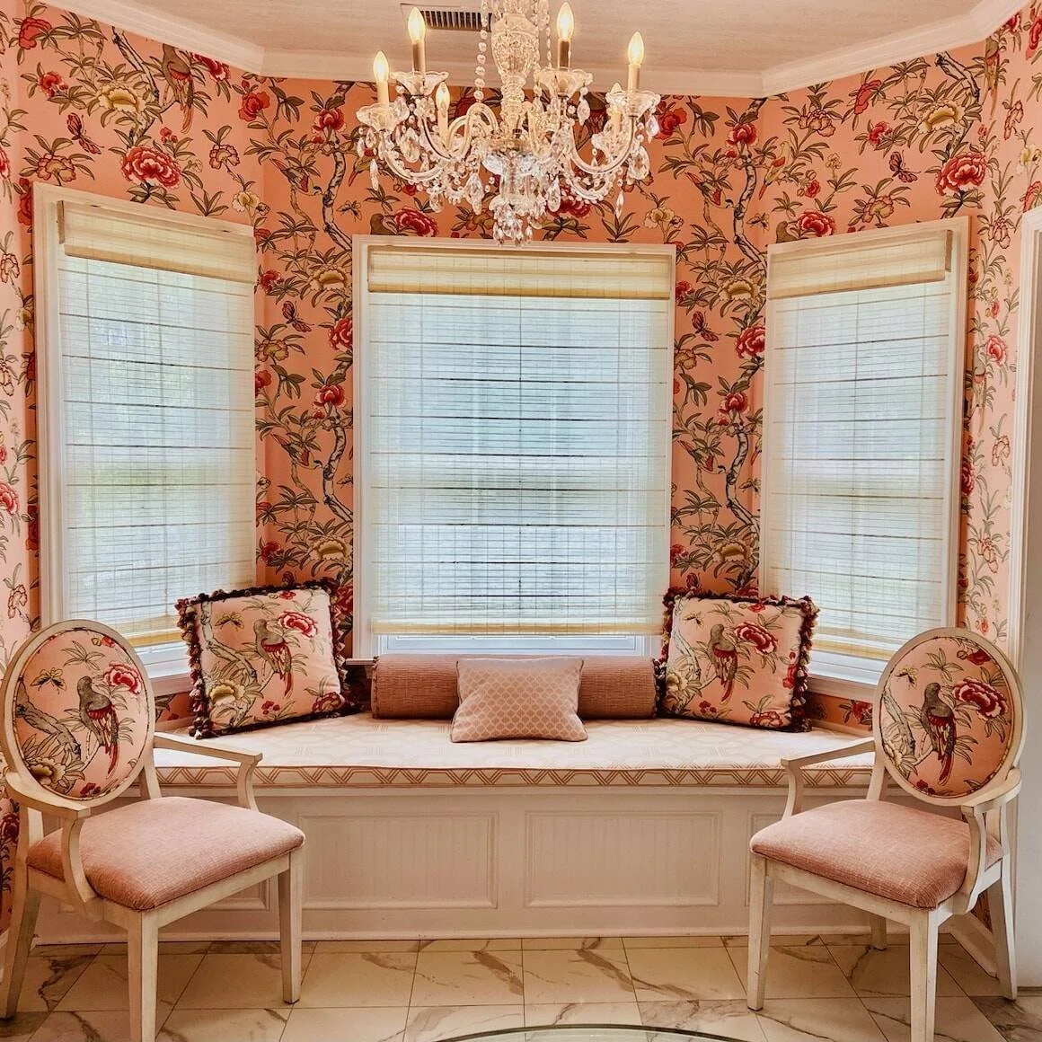 Bay window seat with coral floral wallpaper, matching pillows, and a crystal chandelier designed by Bonnie Boyette Designs