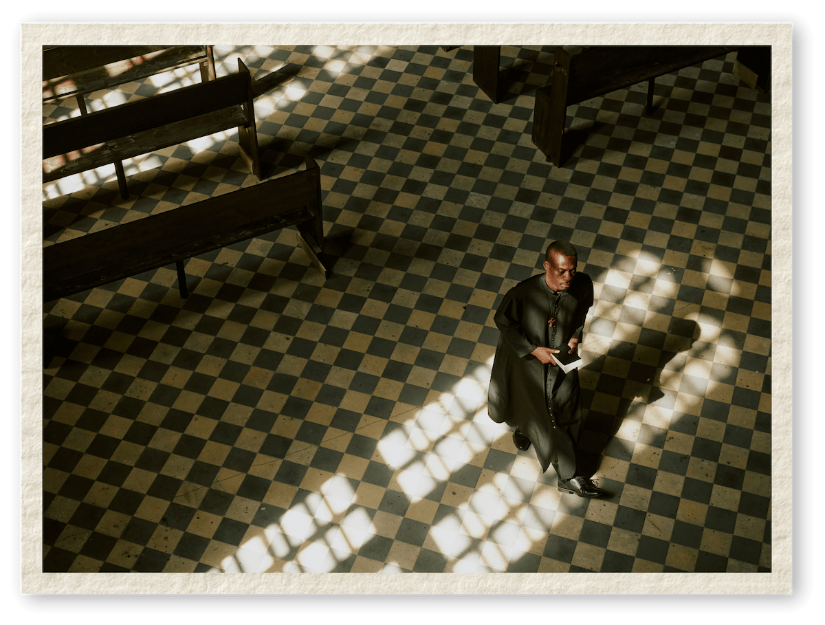 A man dressed in black robes standing inside a church near a window with patterned glass, holding a book or card, illuminated by sunlight filtering through the window.