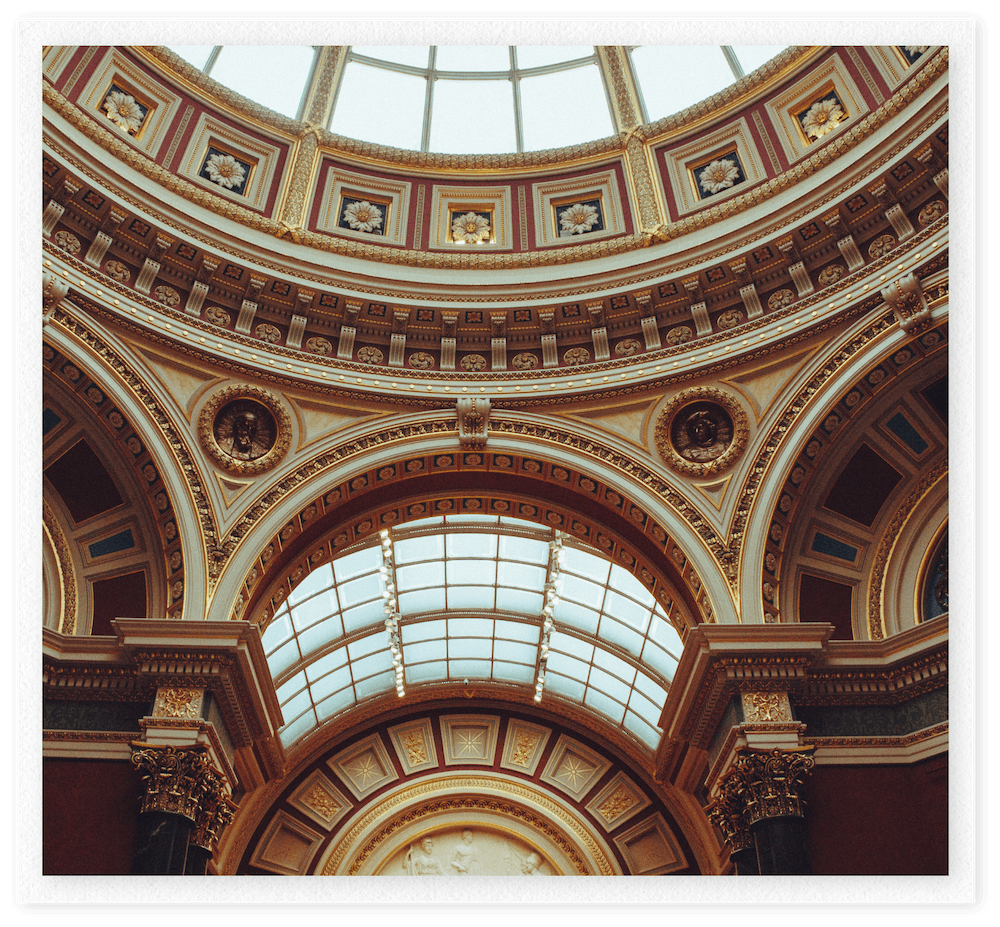 Interior view of an ornate domed ceiling with gold accents, decorative moldings, and a glass skylight at the top, featuring arches and classical details.