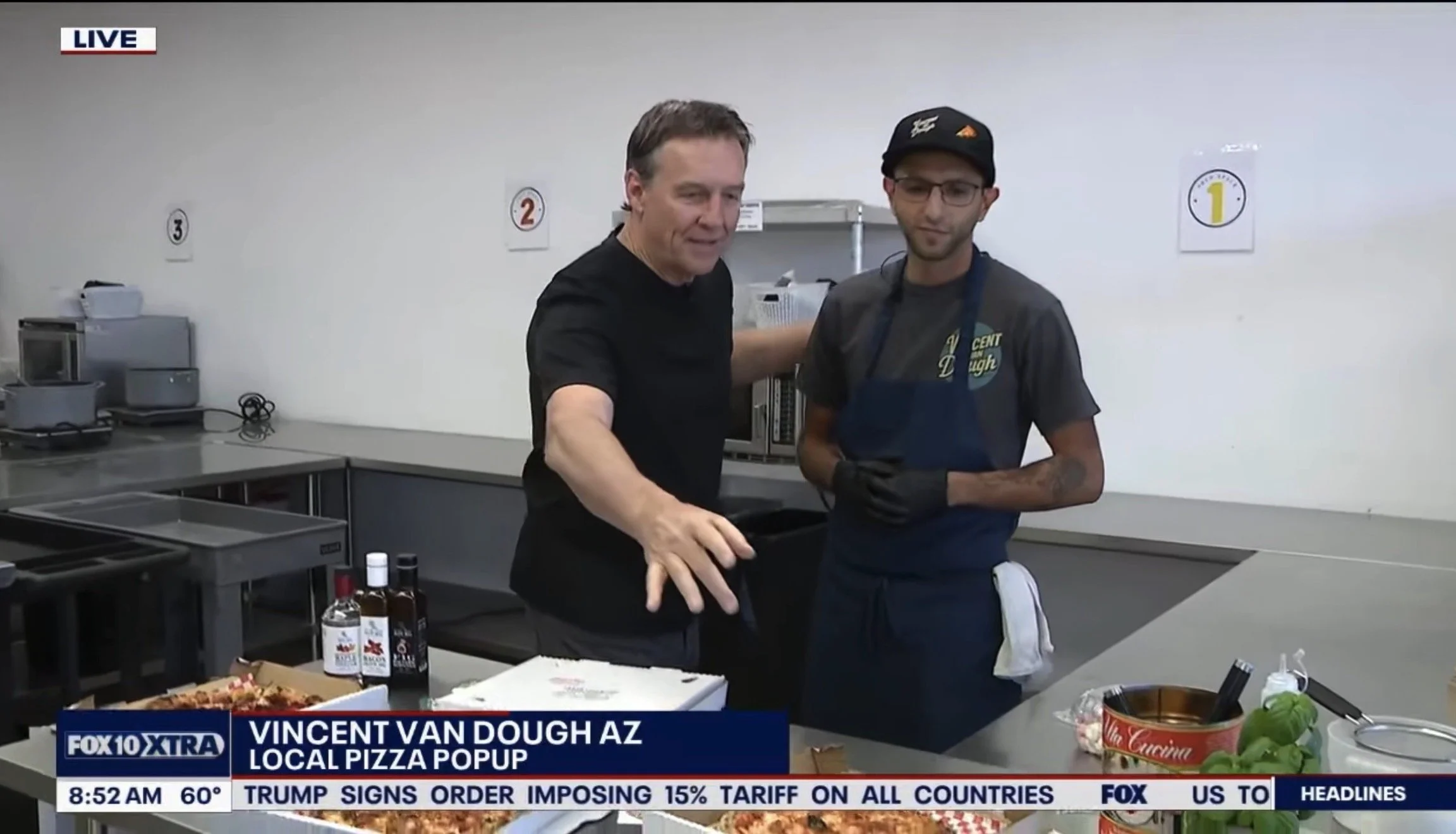 Two men, one in a black shirt and another in a gray shirt with a blue apron, are standing in a kitchen setting, possibly filming a cooking segment. The background shows shelves with kitchen equipment, and there is pizza and bottles of alcohol on the countertop. The scene is from a live Fox 10 TV broadcast about a local pizza popup in Phoenix, Arizona.