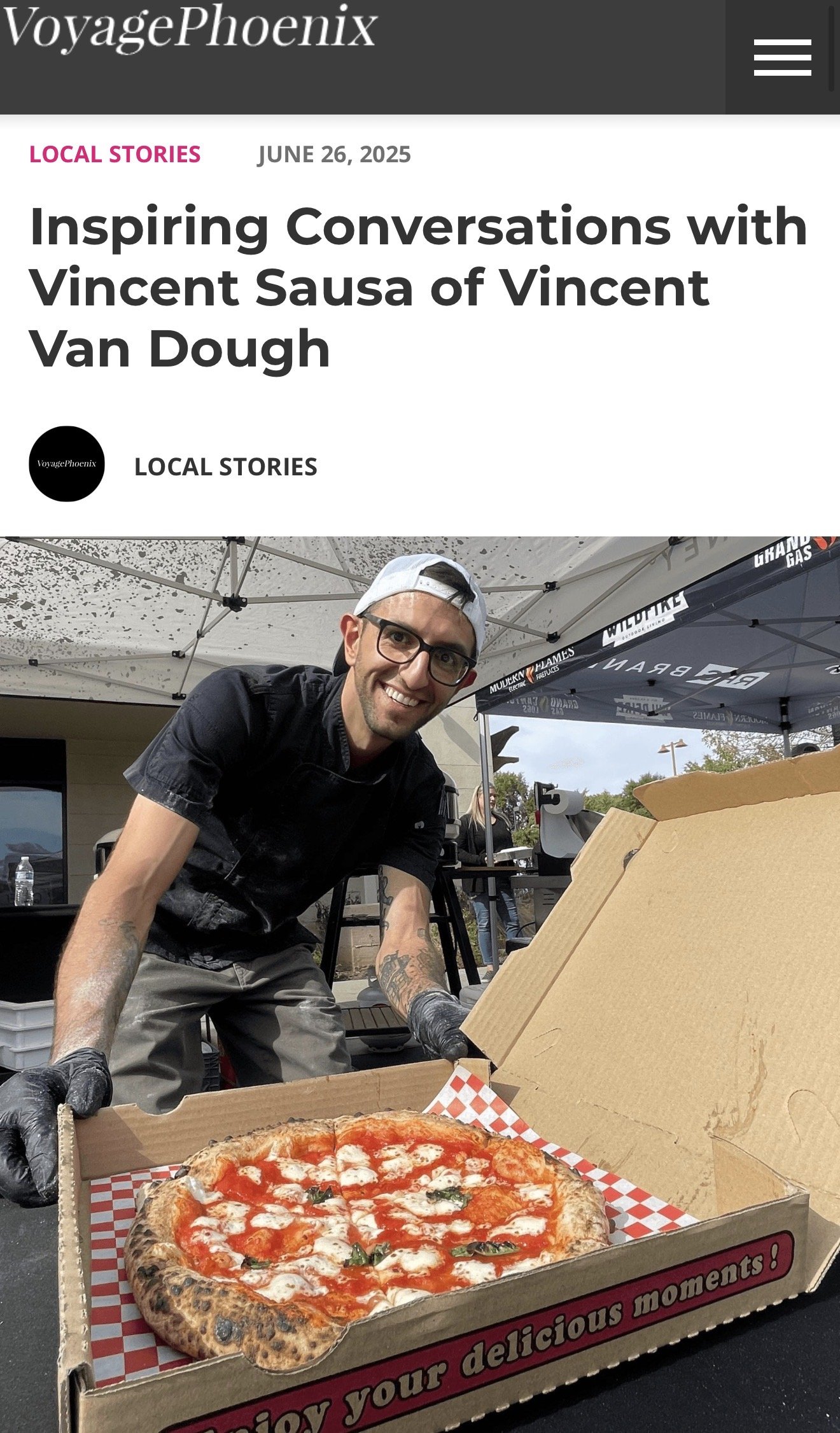 A man wearing glasses and a baseball cap is smiling as he opens a pizza box containing a freshly baked pizza with cheese, tomato sauce, and basil leaves. The scene is outdoors with tents in the background.