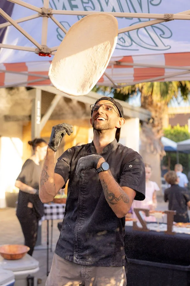 A man in black shirt and glasses tossing pizza dough in the air at an outdoor event.