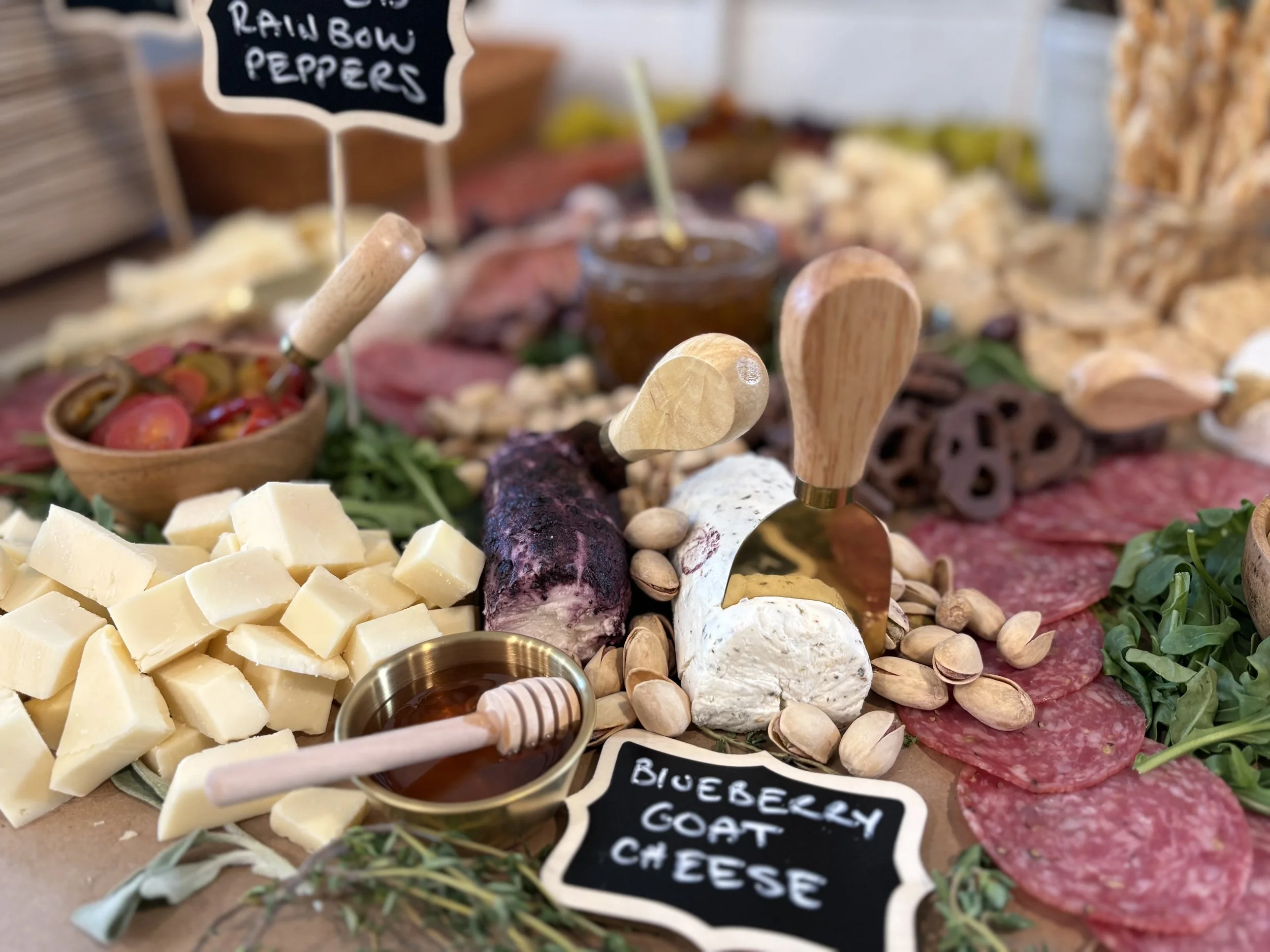 A cheese and charcuterie platter with various cheeses, meats, nuts, and accompaniments, including a small bowl of honey and honey dipper, garnished with greenery and labeled with small chalkboard signs.