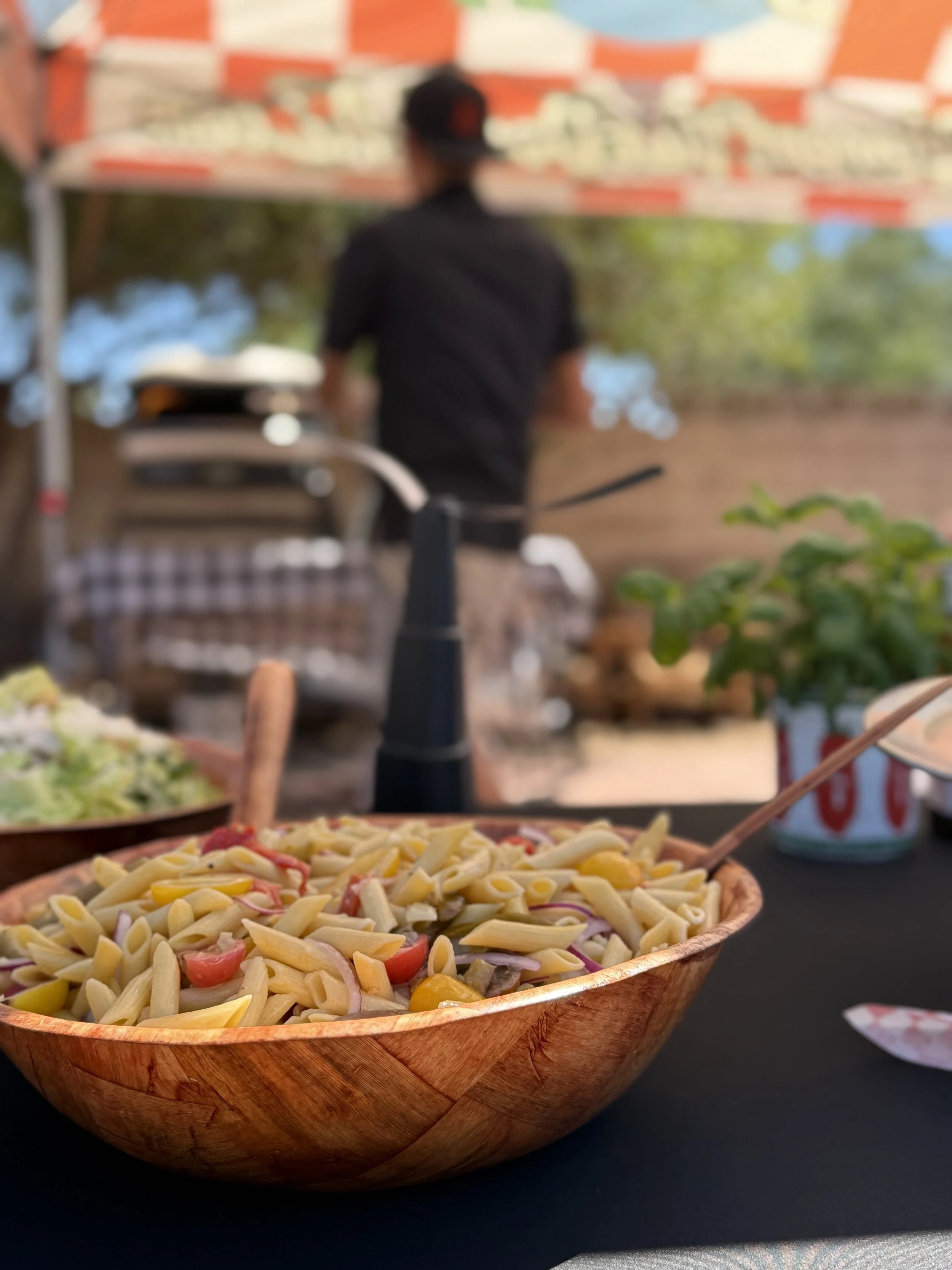 A bowl of pasta salad with cherry tomatoes, onions, and shredded cheese on a black table with a can of soda and outdoor trees in the background.