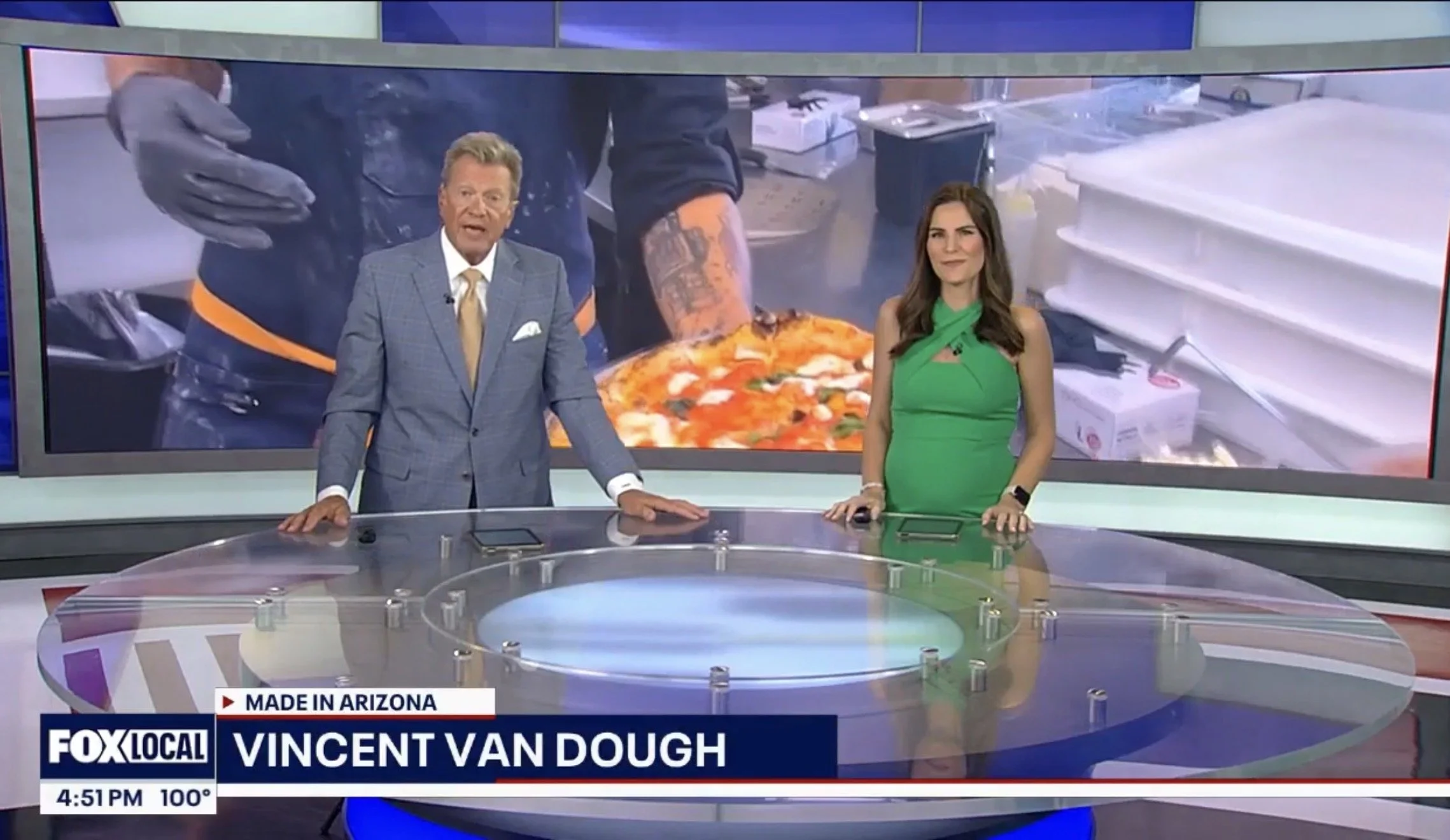 News anchors presenting on a television set with a large screen showing a pizza being made in the background. The male anchor wears a gray plaid suit and a gold tie, and the female anchor wears a green dress. Information on the screen indicates it's 4:51 PM and the temperature is 100°F.
