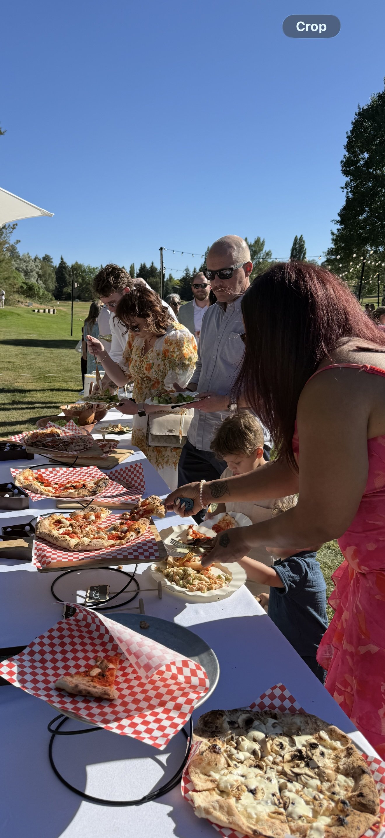 People at an outdoor buffet table selecting pizza slices on a sunny day with clear blue sky, trees, and string lights in the background.